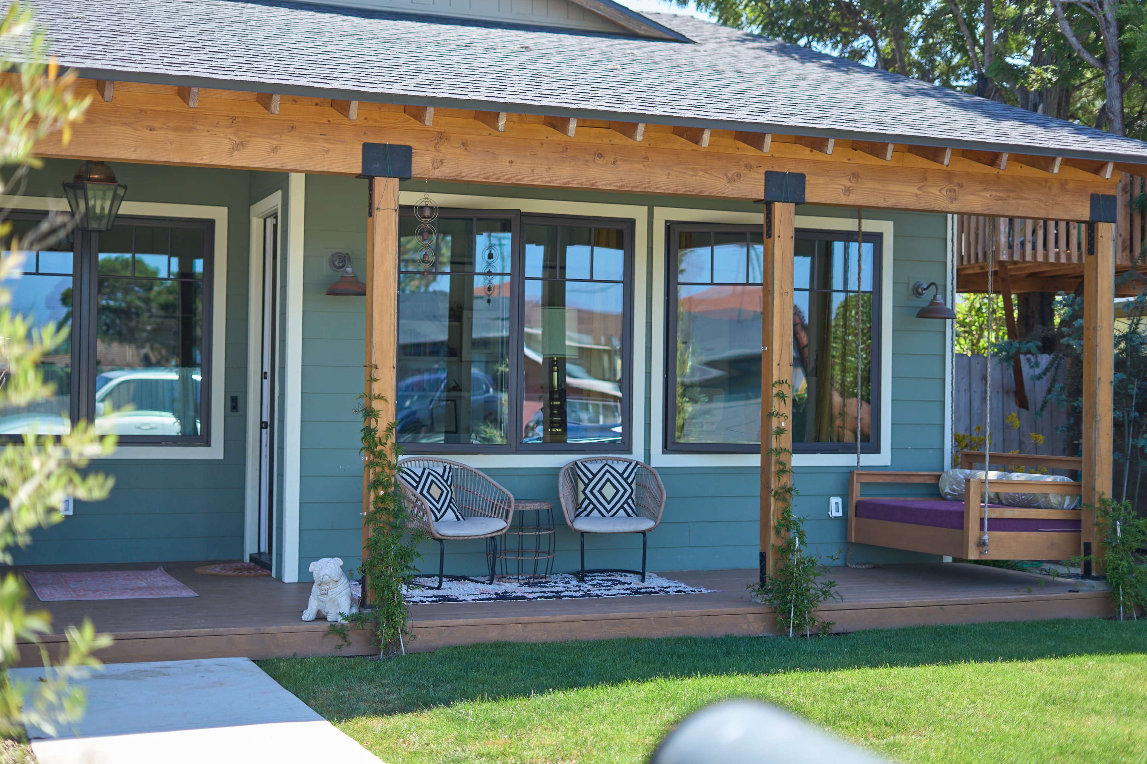 The image depicts a modern house with a covered porch, featuring two chairs and a small white dog on the wooden deck.