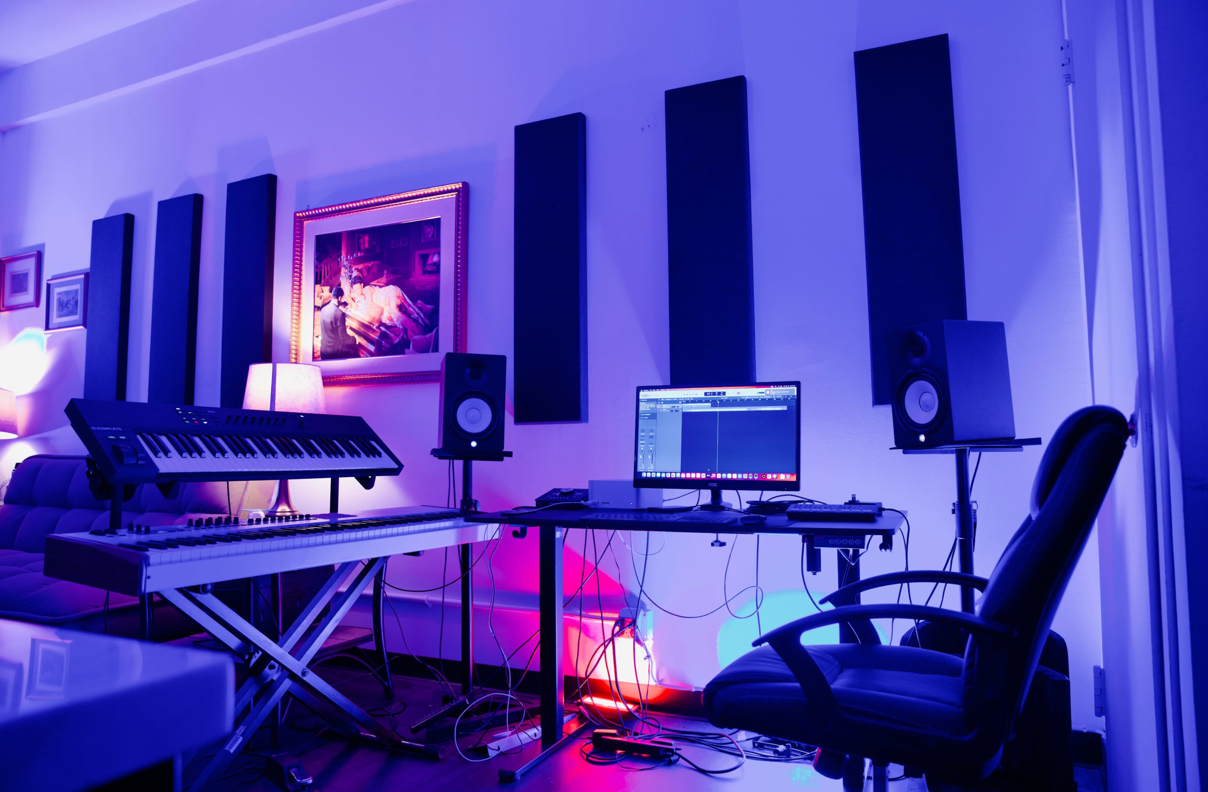 The image shows a music studio setup with a keyboard, computer, studio monitors, and acoustic panels, illuminated by colored lights.