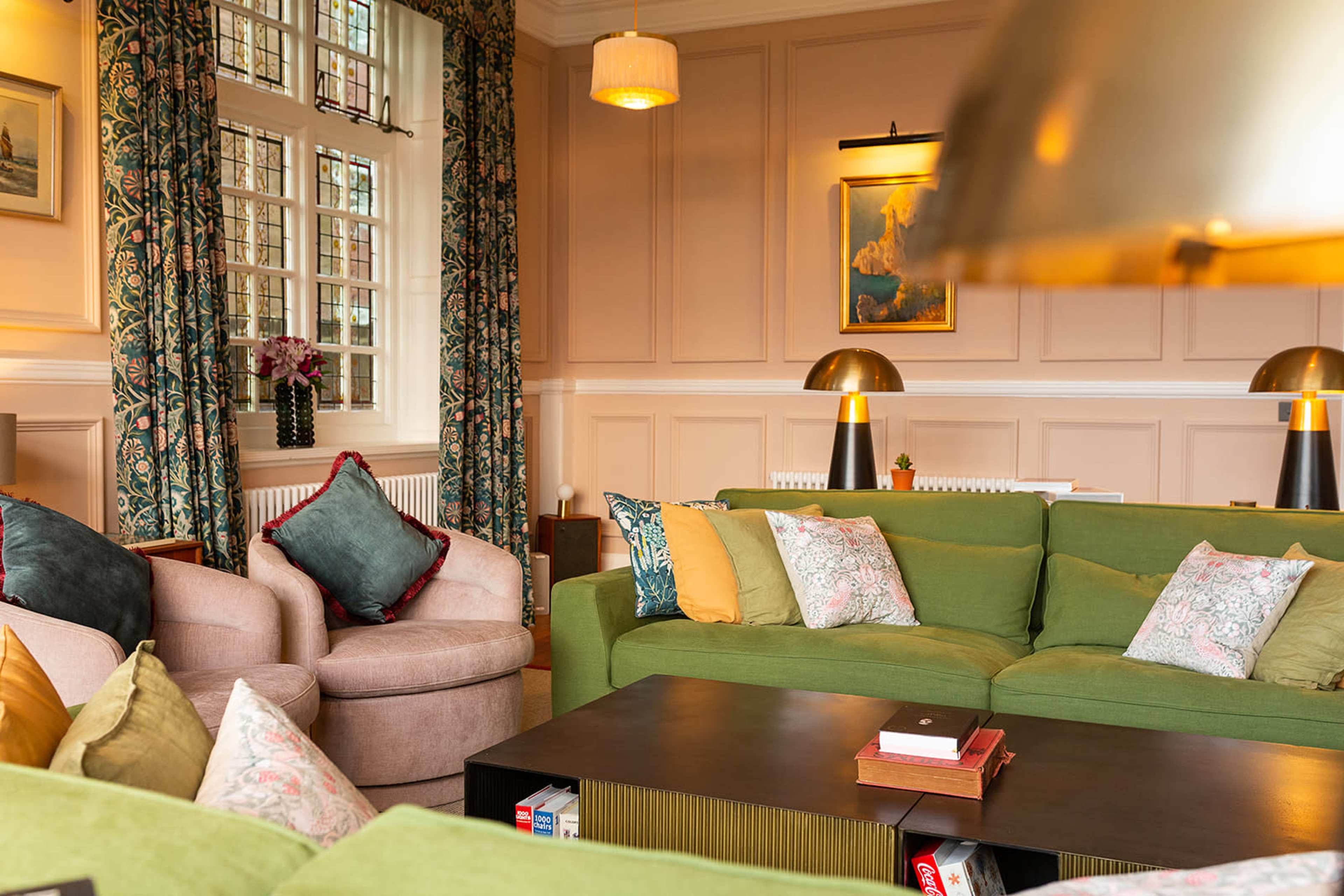 The scene depicts a cozy living room with a green sectional sofa, two armchairs, and a dark coffee table, illuminated by soft lighting and adorned with decorative pillows.