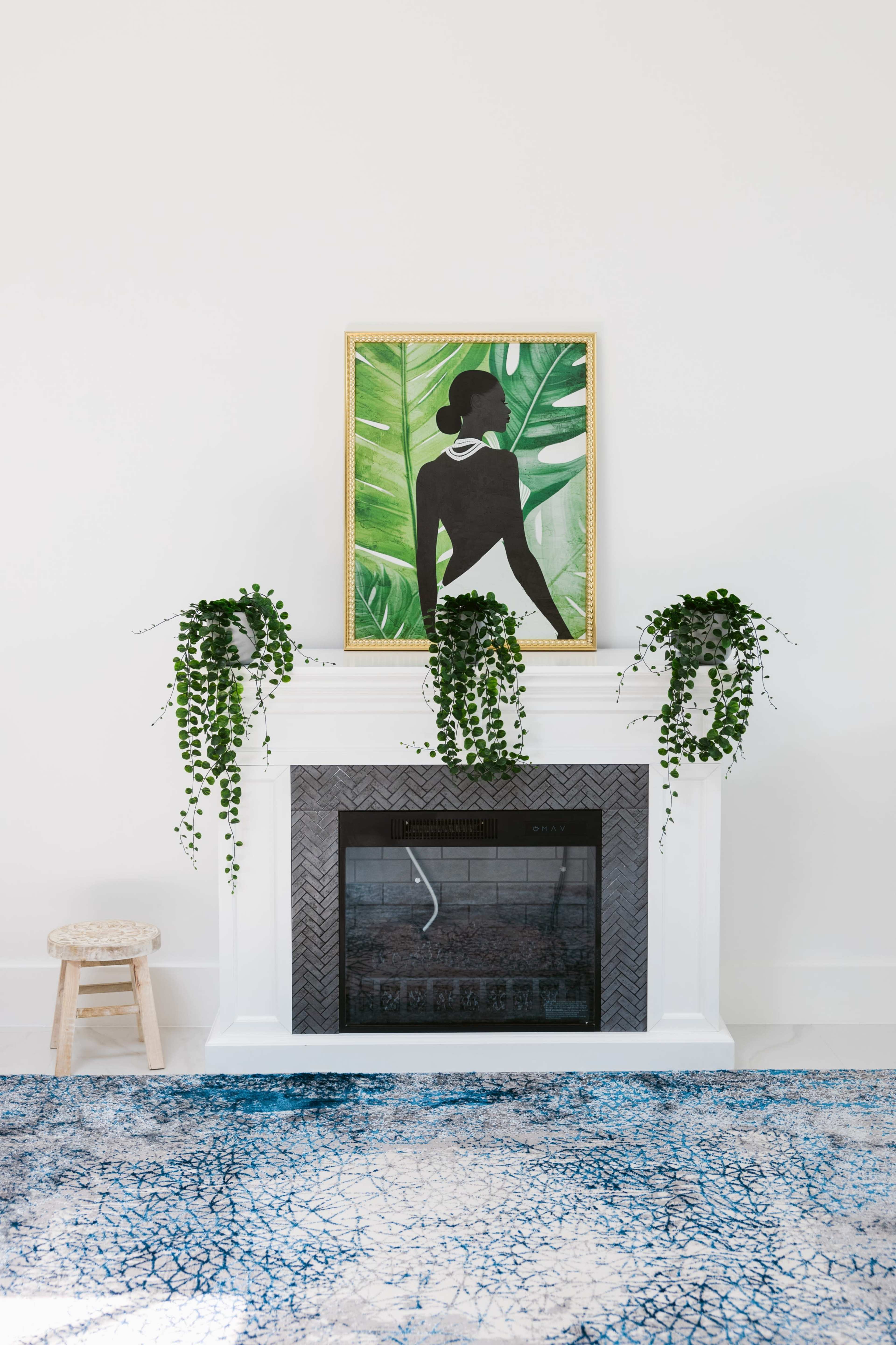 The image features a white fireplace adorned with potted plants on either side, a colorful artwork above, and a patterned rug in front.