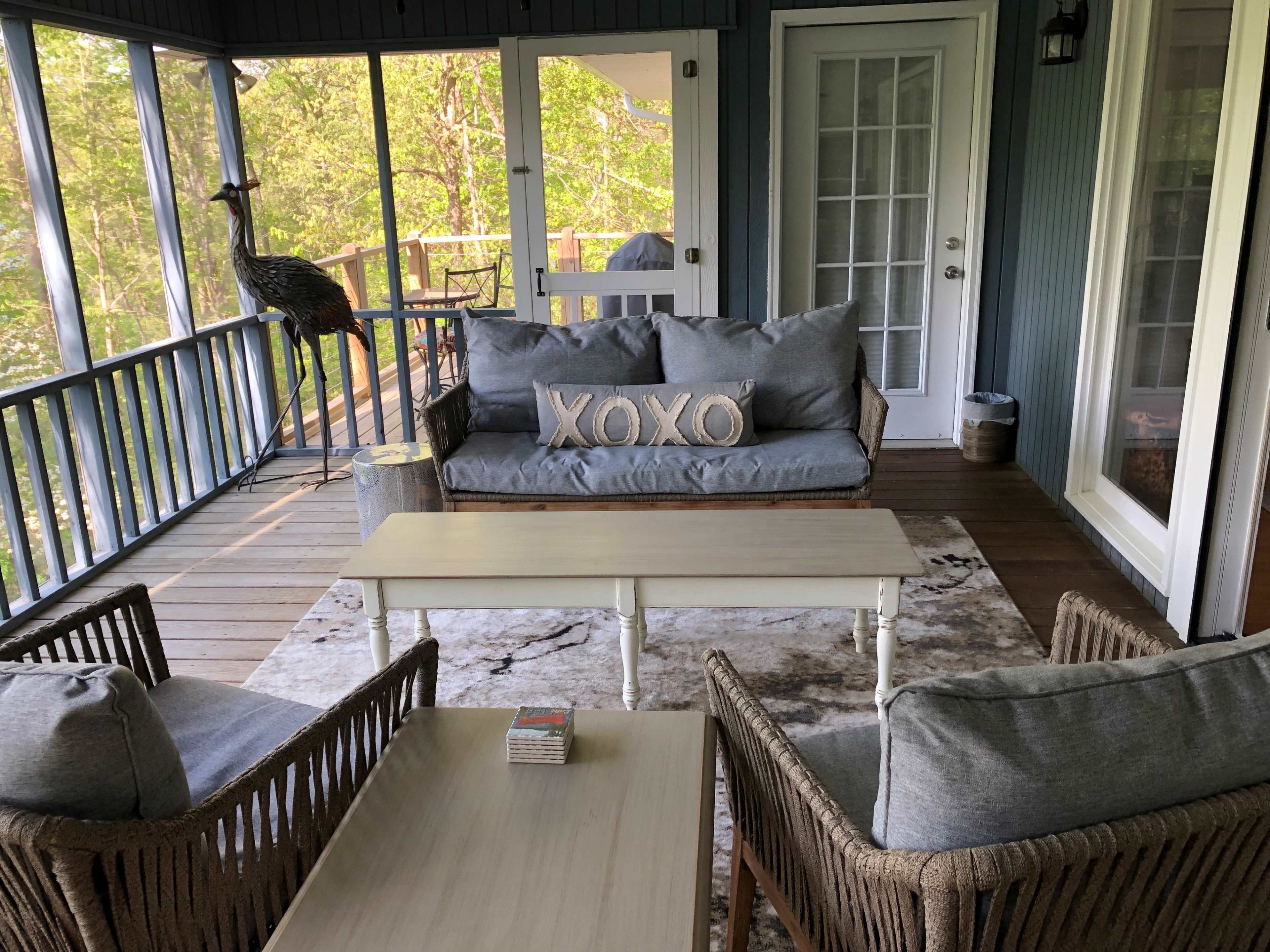 A screened-in porch with a gray sofa, two wicker chairs, and a small wooden coffee table, all arranged around a patterned rug.