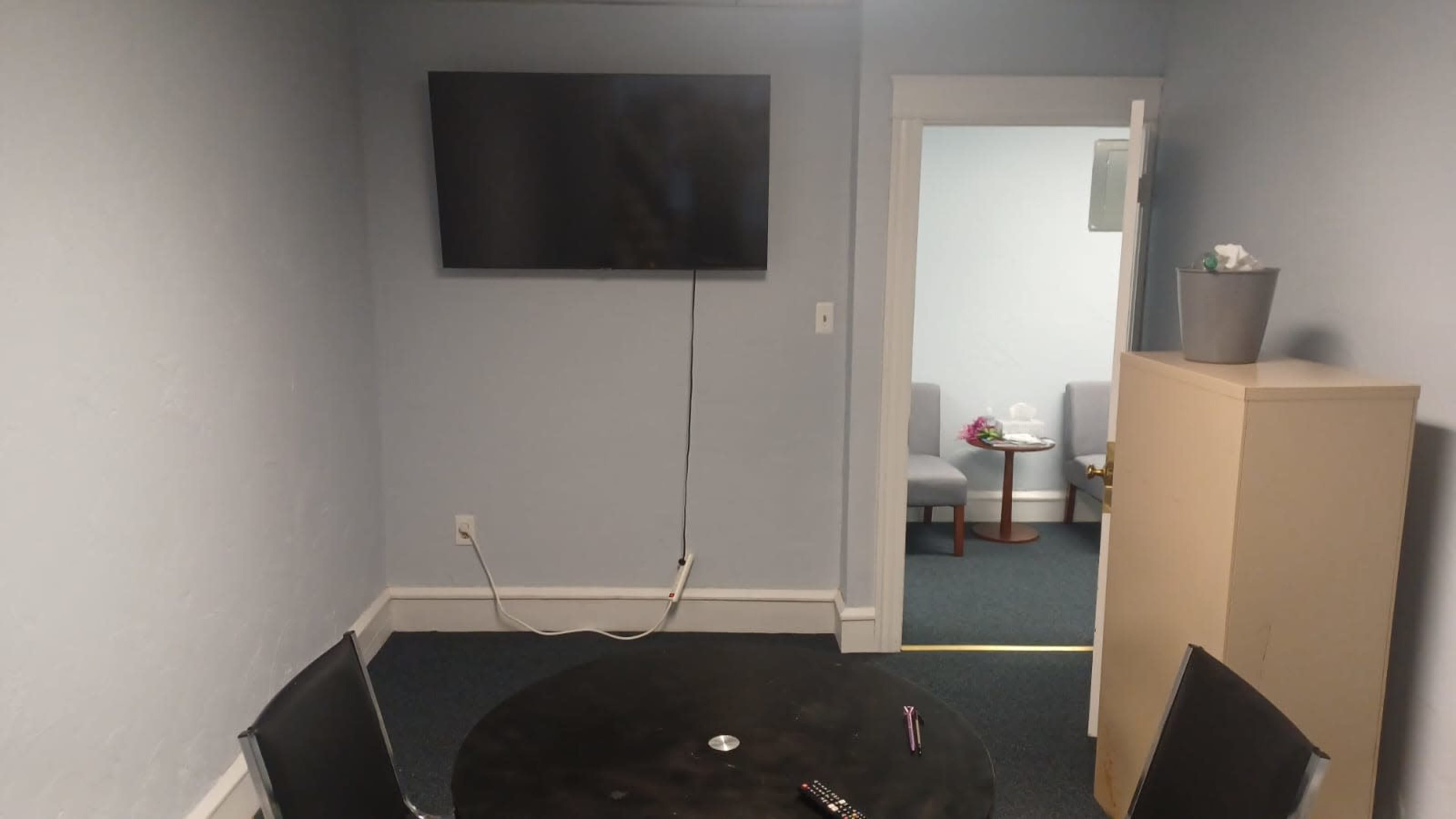 A small room features a black table with two chairs, a wall-mounted TV, and a doorway leading to another room with two seating options.