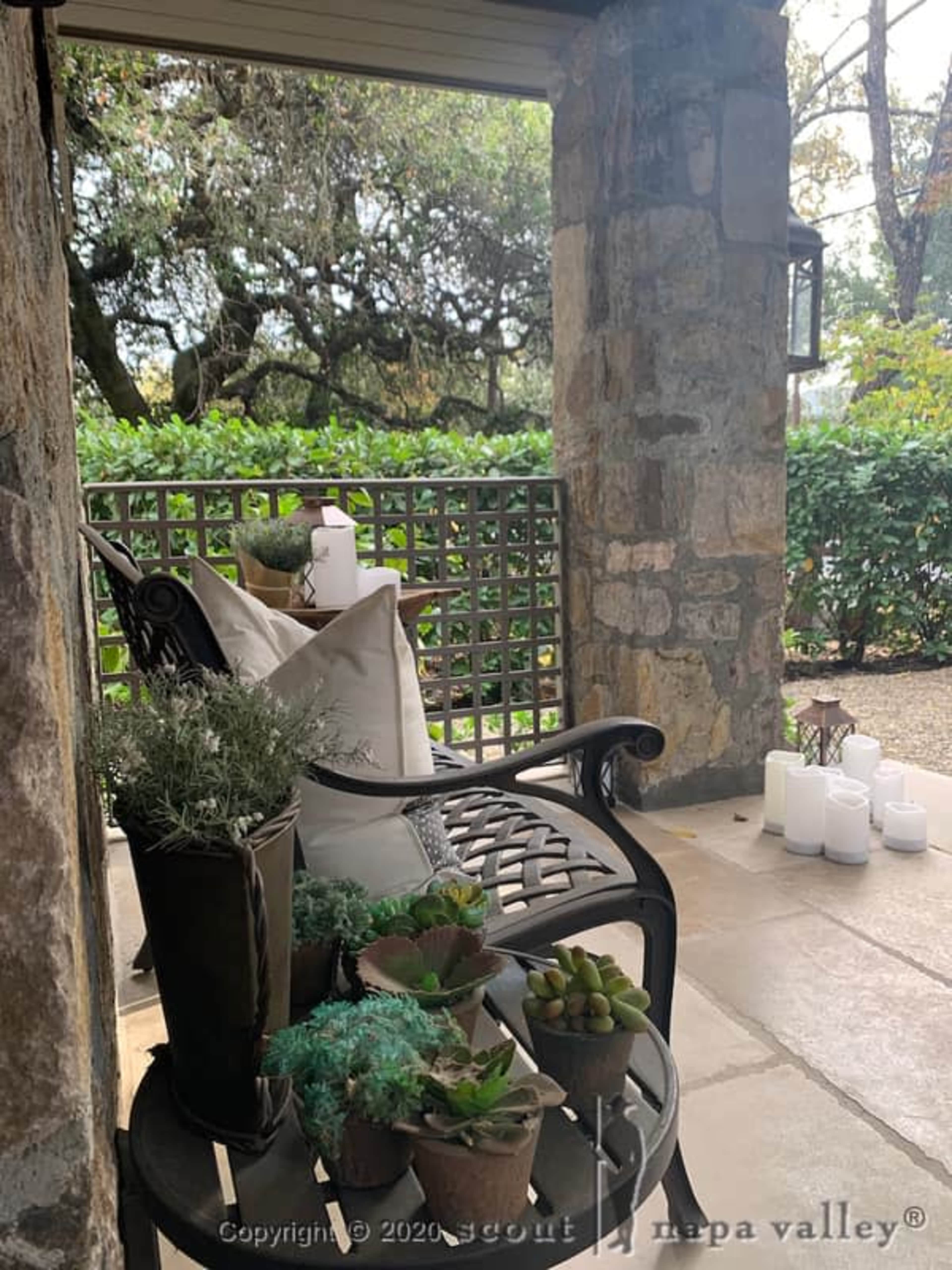 A stone patio features a metal chair with decorative cushions and potted succulents, along with white candles arranged nearby.