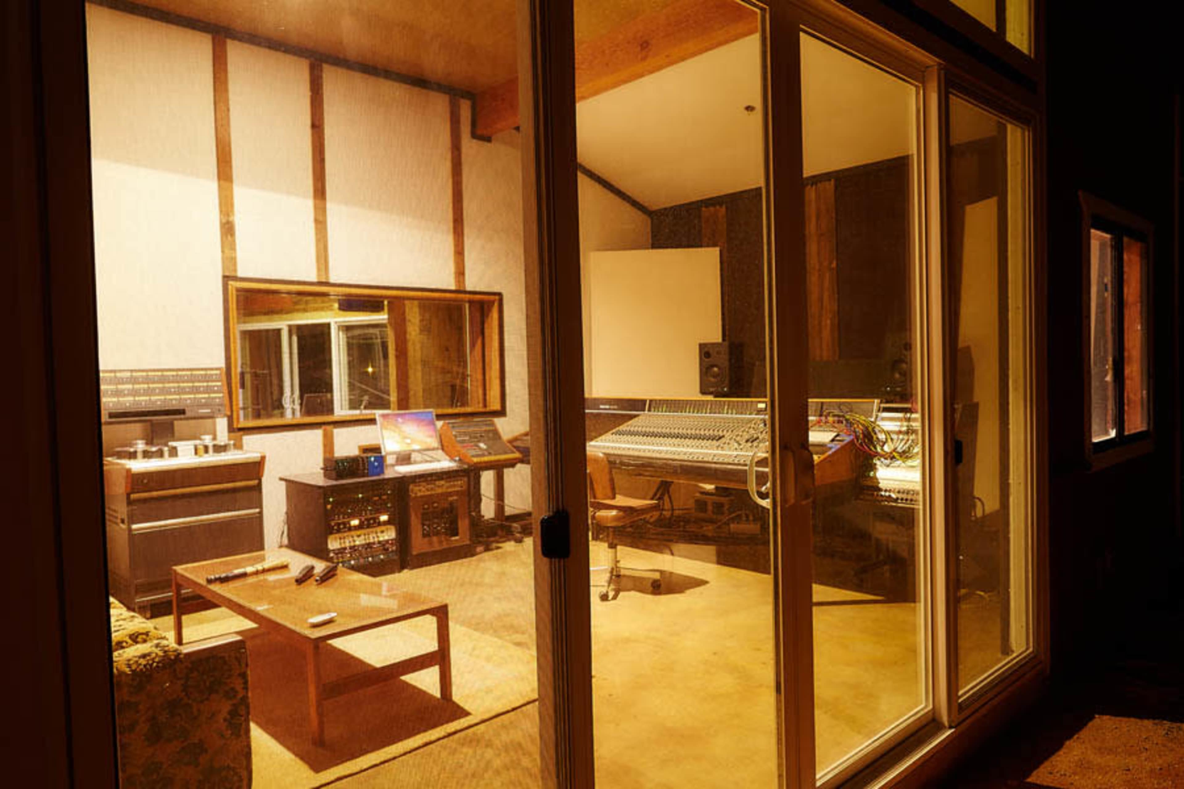 A recording studio is visible through large glass windows, featuring a sound desk, monitors, and various audio equipment.