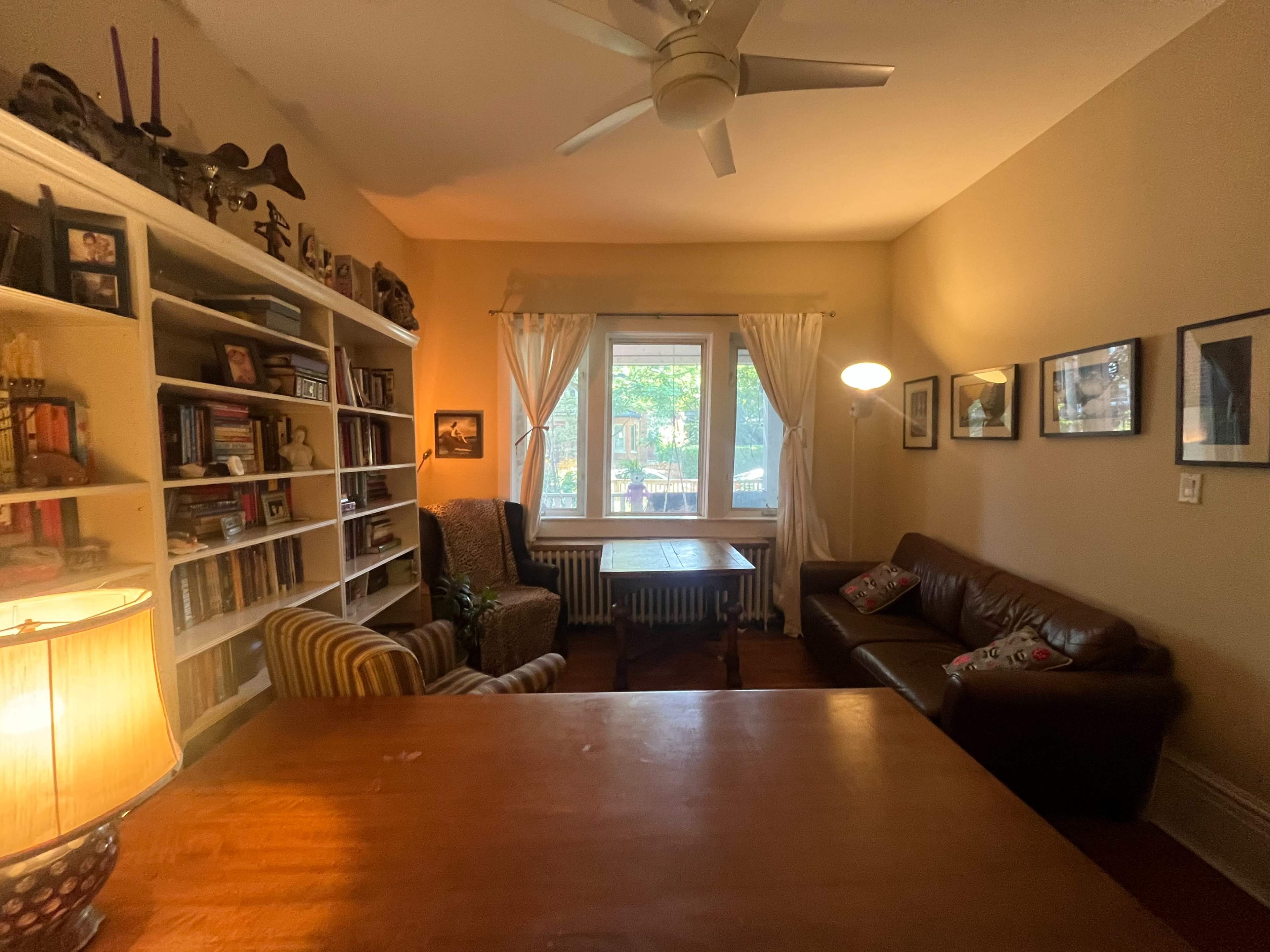 The image shows a cozy living room with a wooden table at the center, surrounded by a bookshelf, a sofa, and a chair, with natural light coming through a window.