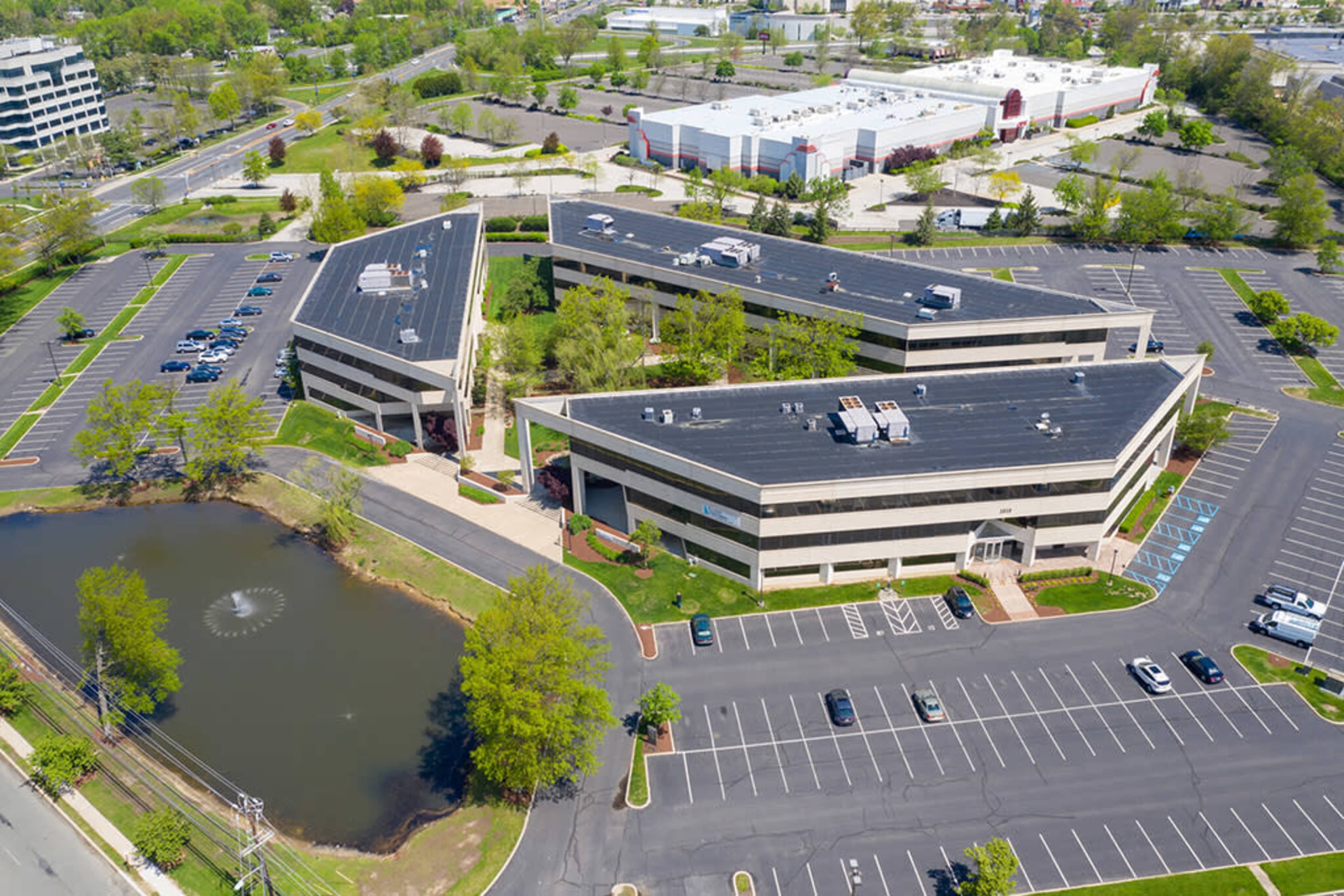 The image shows an aerial view of a commercial complex with modern buildings, surrounded by well-maintained landscaping and a pond, along with a spacious parking lot that appears mostly empty.