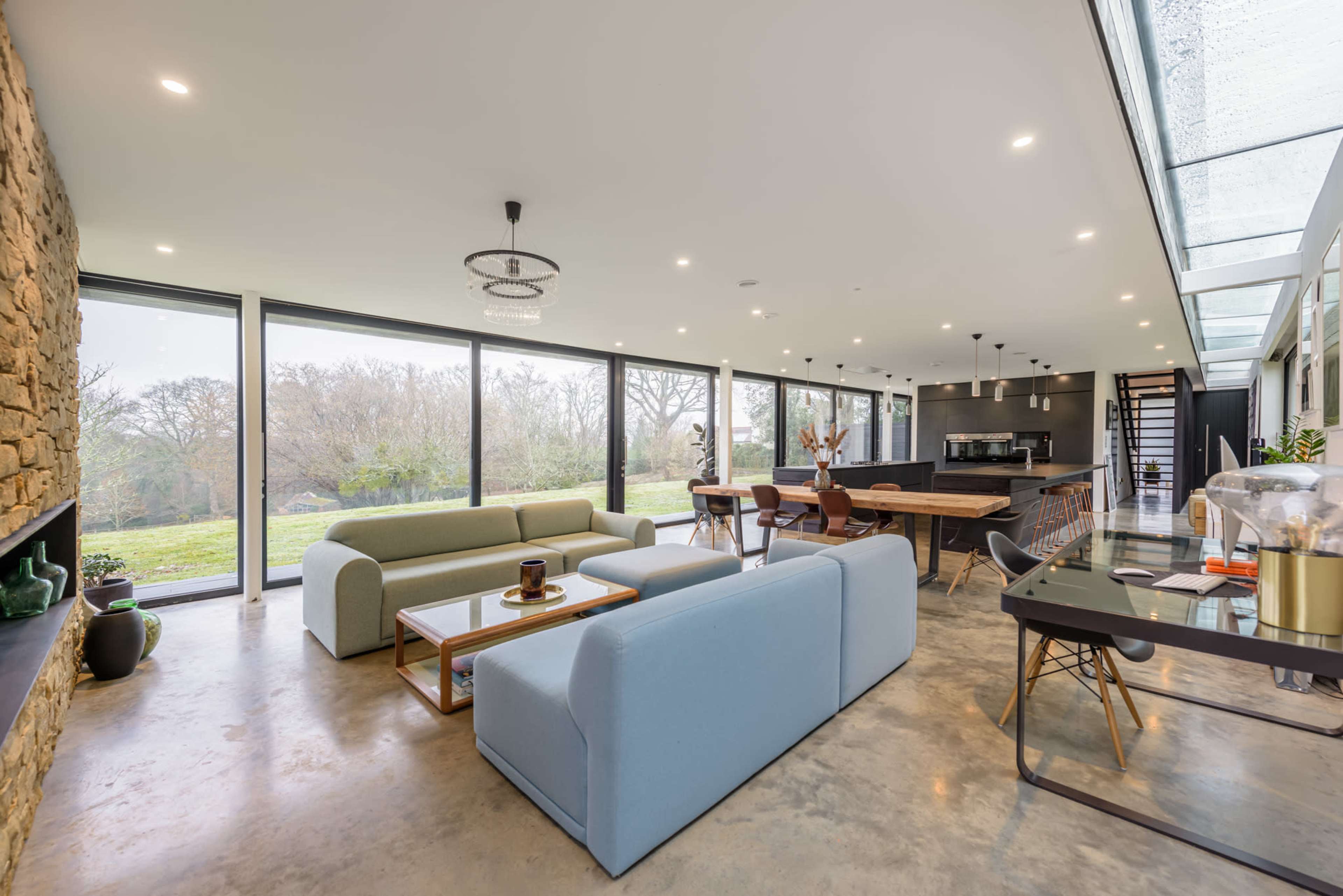 A modern open-plan living space with large windows, a dining area, and contemporary furnishings, including a blue sofa and a wooden dining table.