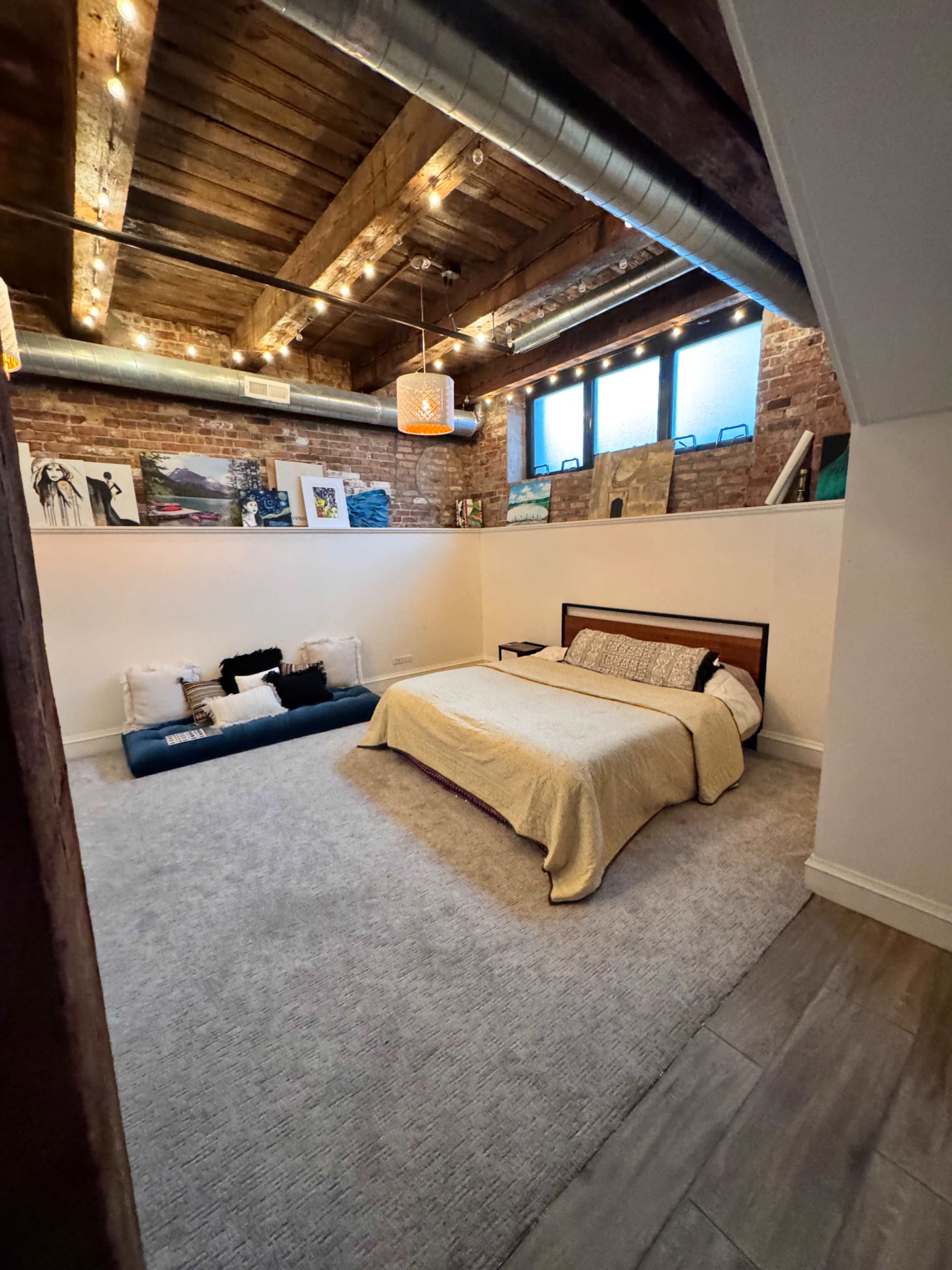 The image shows a cozy bedroom with a bed, decorative pillows, exposed brick walls, and wooden beams in the ceiling.