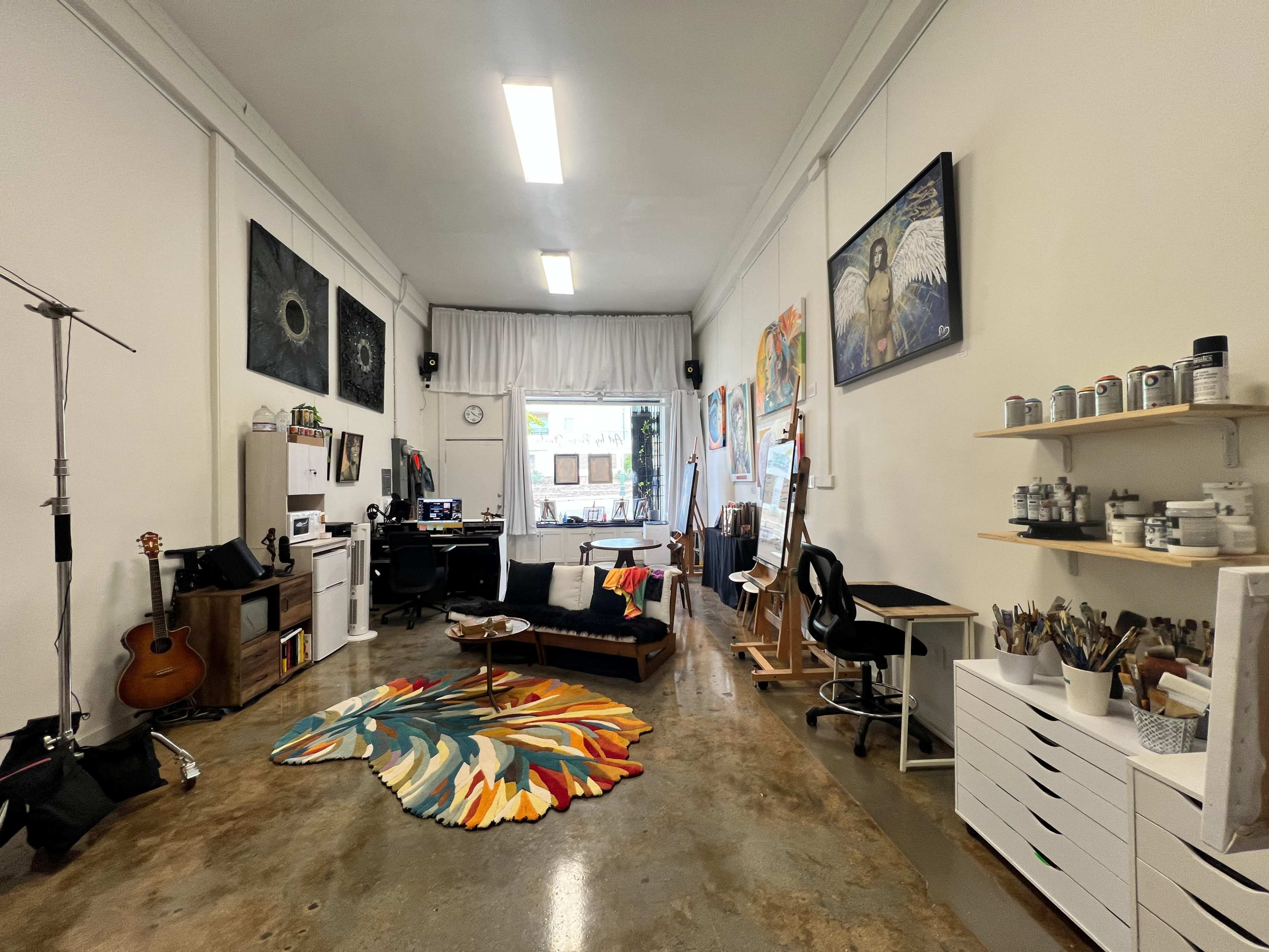 The image shows an artist's studio featuring an eclectic arrangement of artwork, furniture, and art supplies, with natural light coming in through a large window.