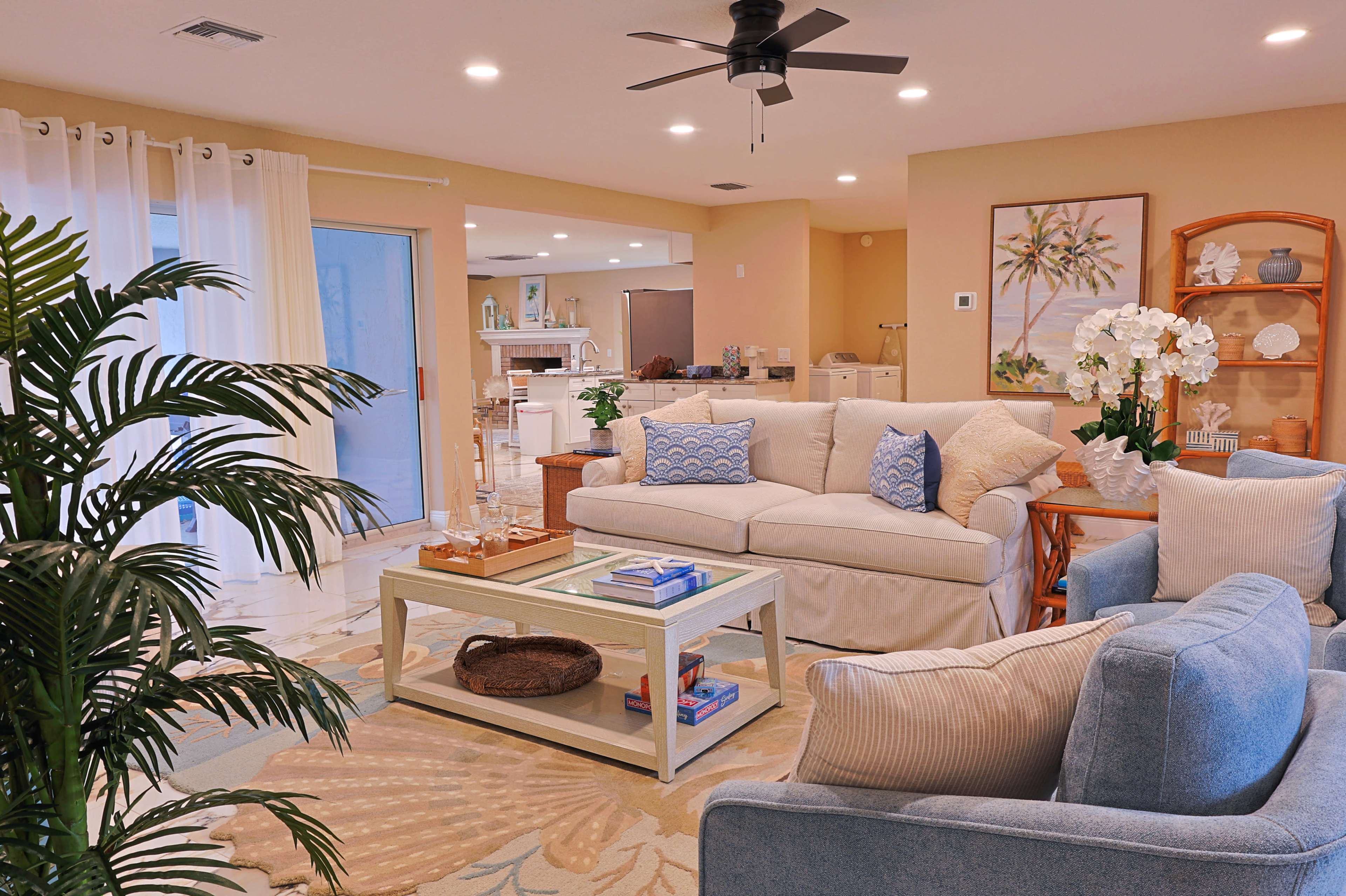 The image shows a brightly lit living room with a beige sofa, blue accent chairs, a glass coffee table, and a tropical-themed area rug.