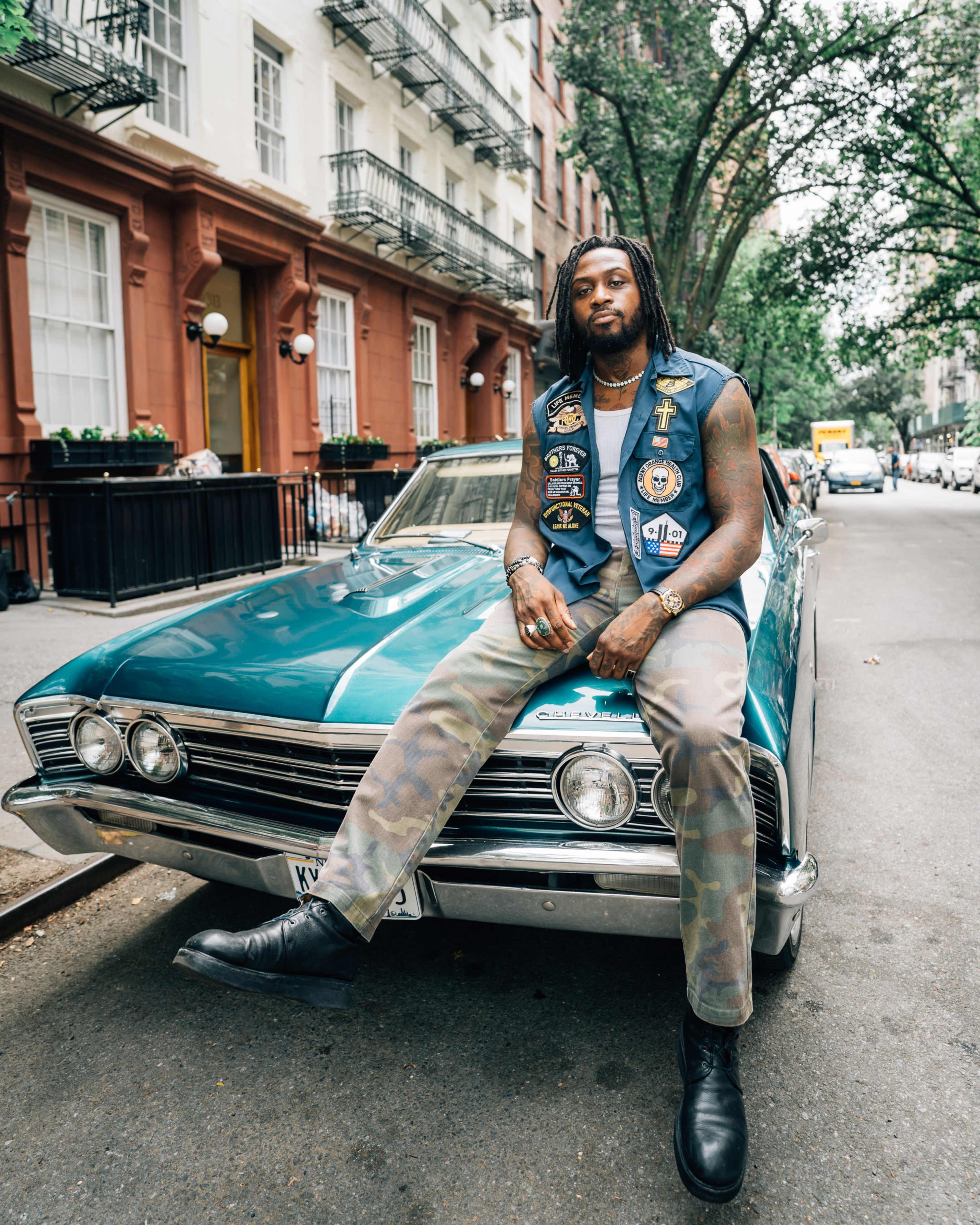A man in a sleeveless jacket adorned with patches sits on the hood of a vintage blue car parked on a city street flanked by apartment buildings.