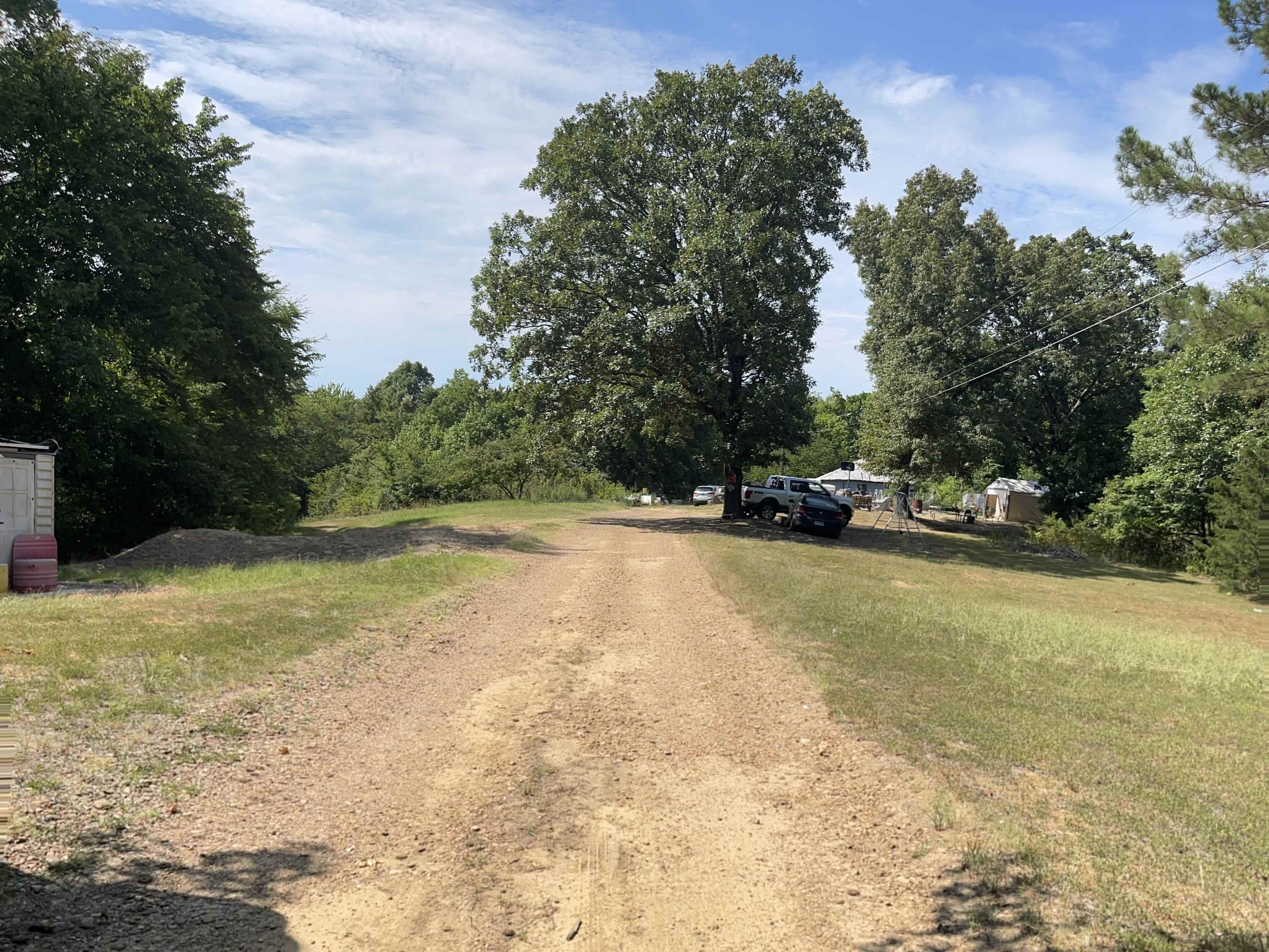 A dirt road leads through a grassy area with trees on either side and vehicles parked in the background.