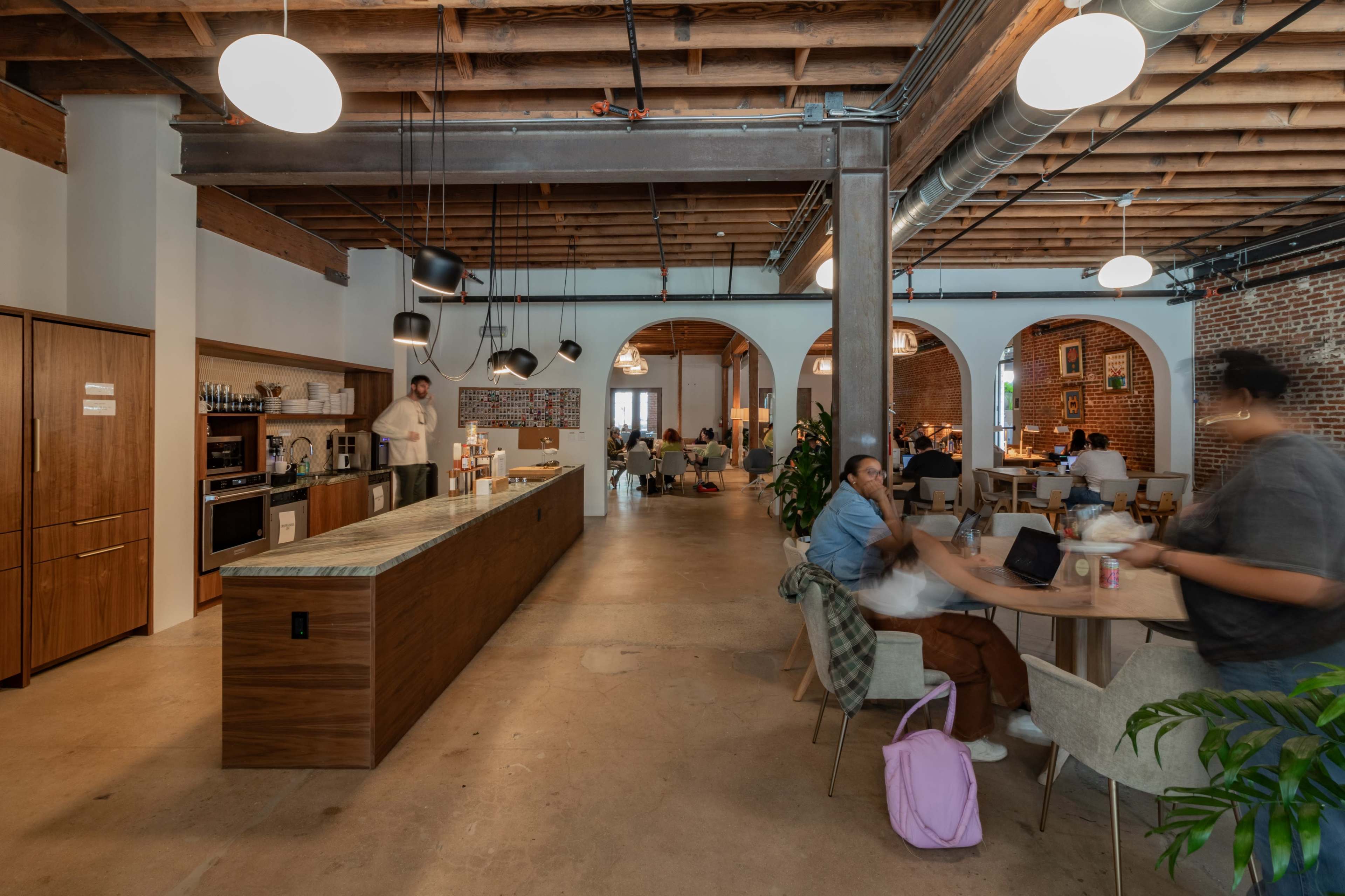 A spacious coworking space with a kitchen area, tables, and people engaged in work and conversations under exposed wooden beams and industrial lighting.