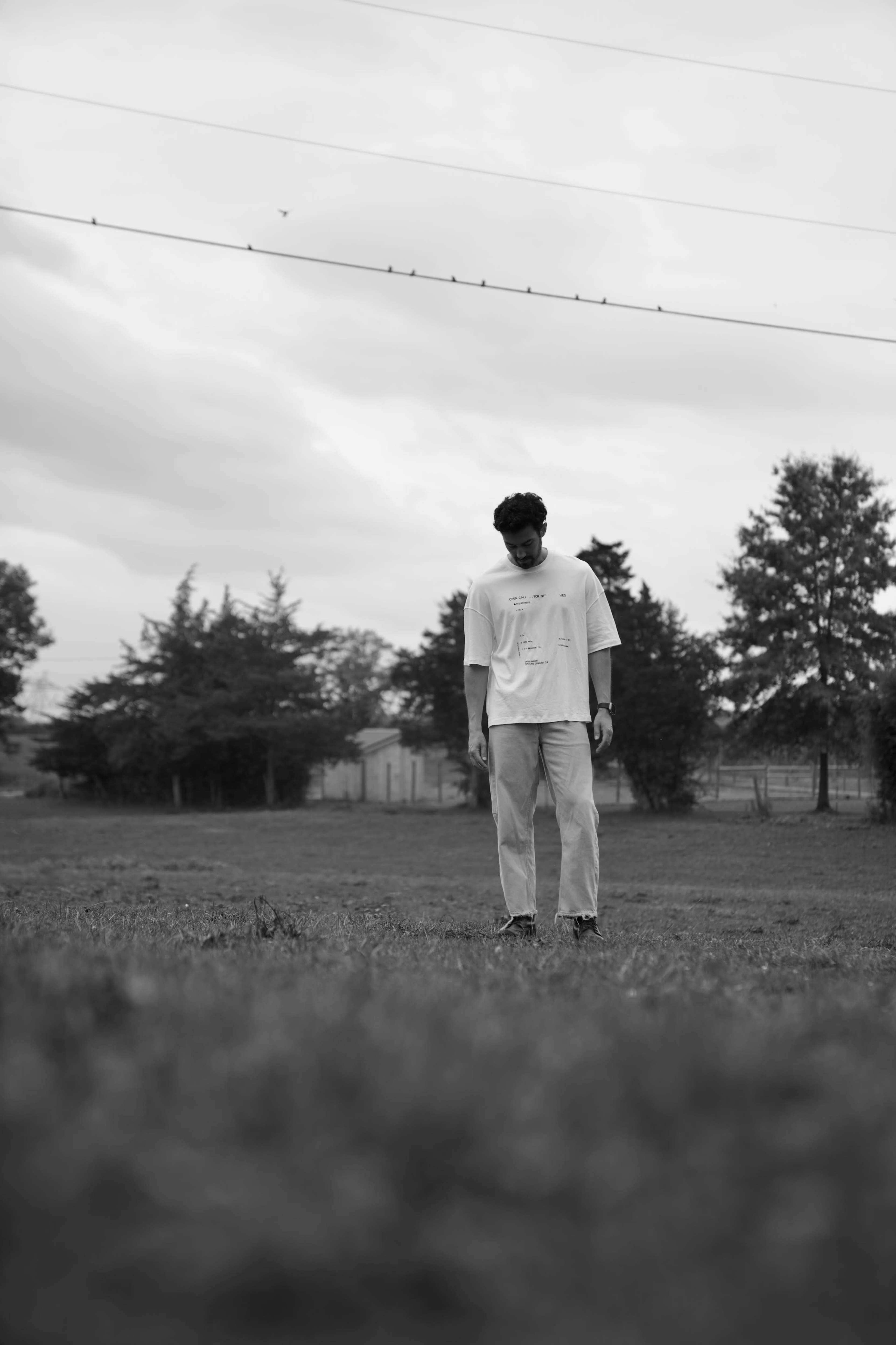 A man stands in a grassy field, looking down, with trees and a cloudy sky in the background.