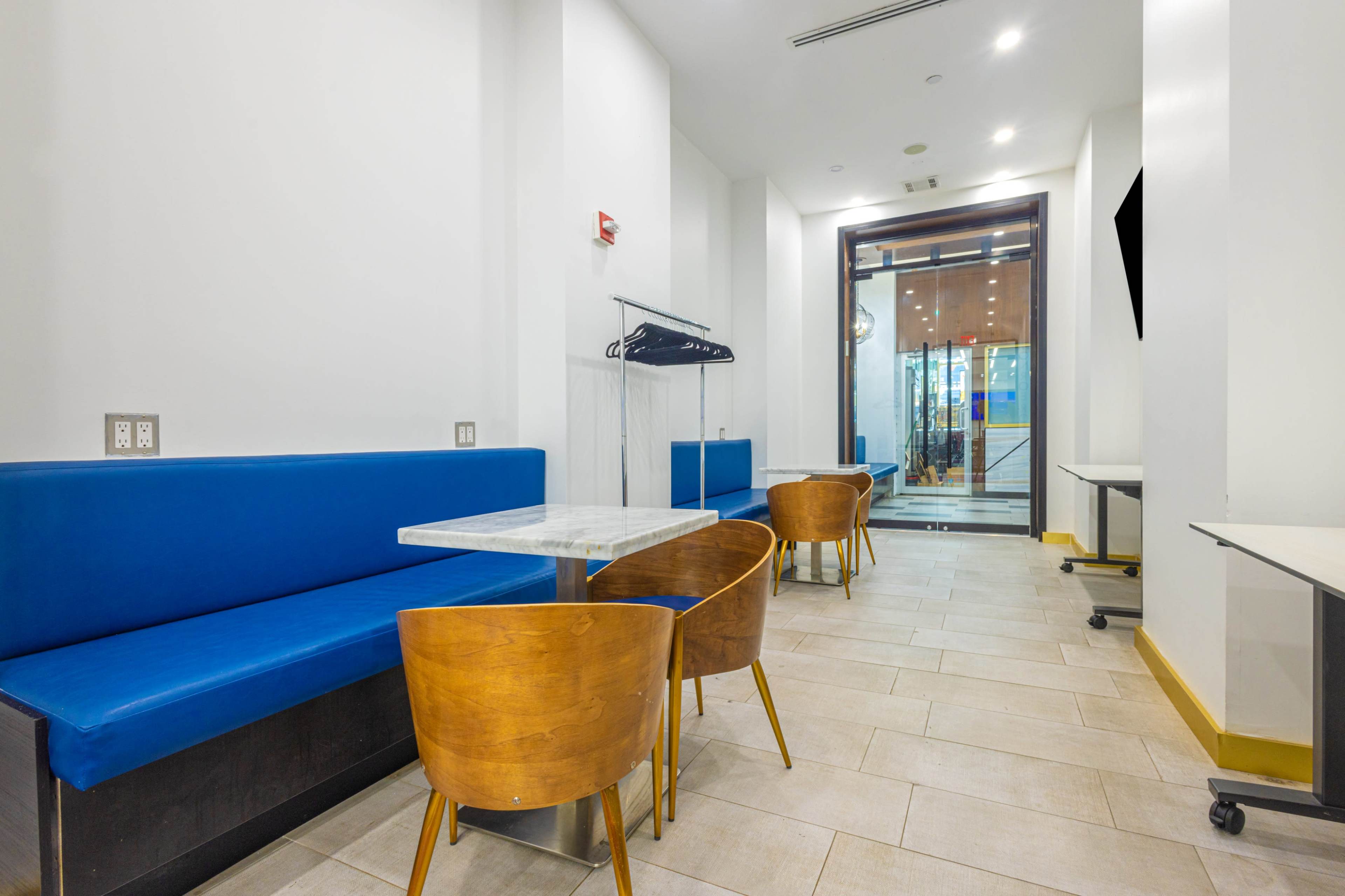 The image shows a modern dining area with blue benches and wooden chairs arranged around marble tables, leading to an entrance with large glass doors.