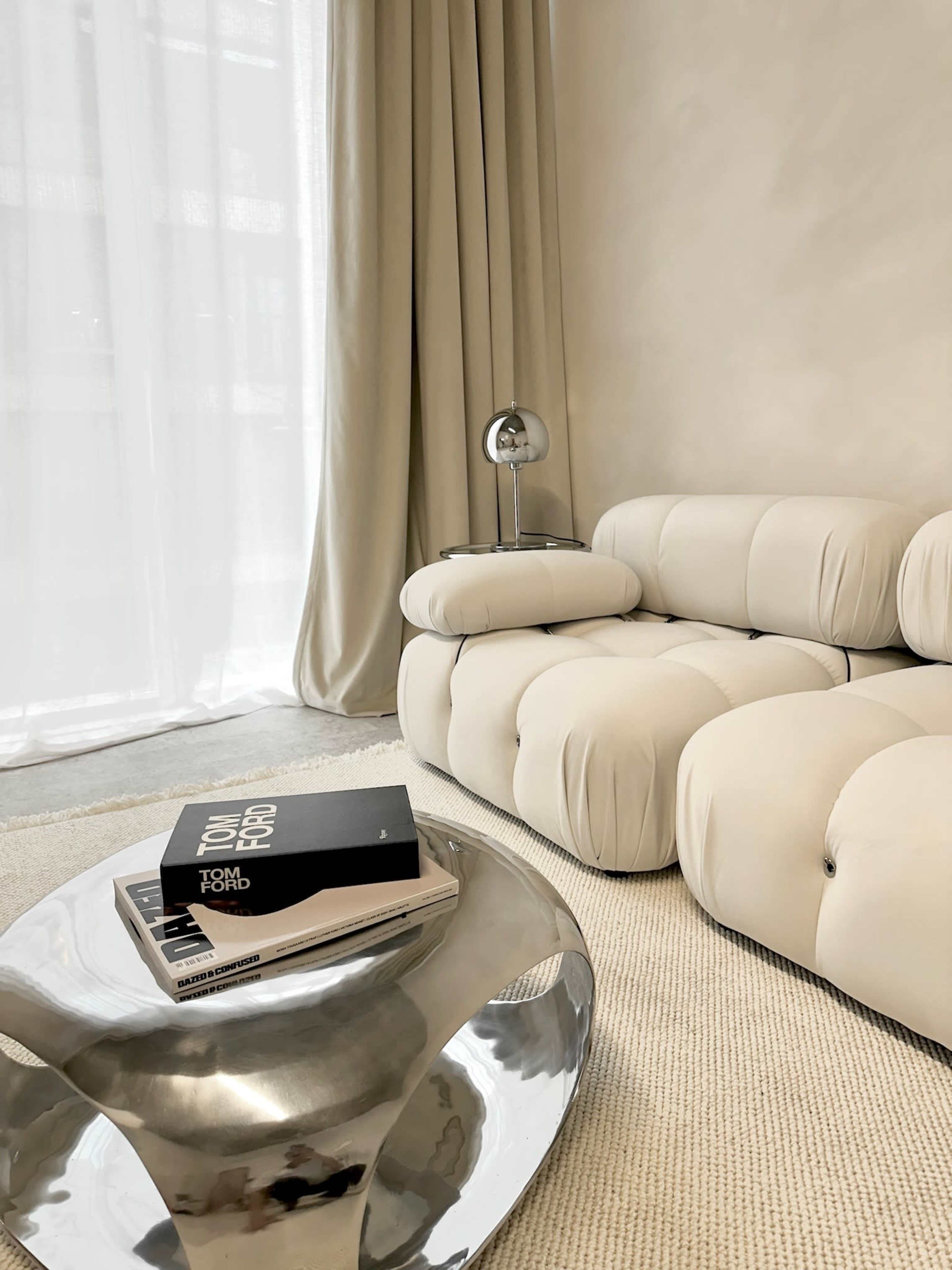 A modern living room features a light-colored, tufted sofa and a reflective coffee table with books placed on top.