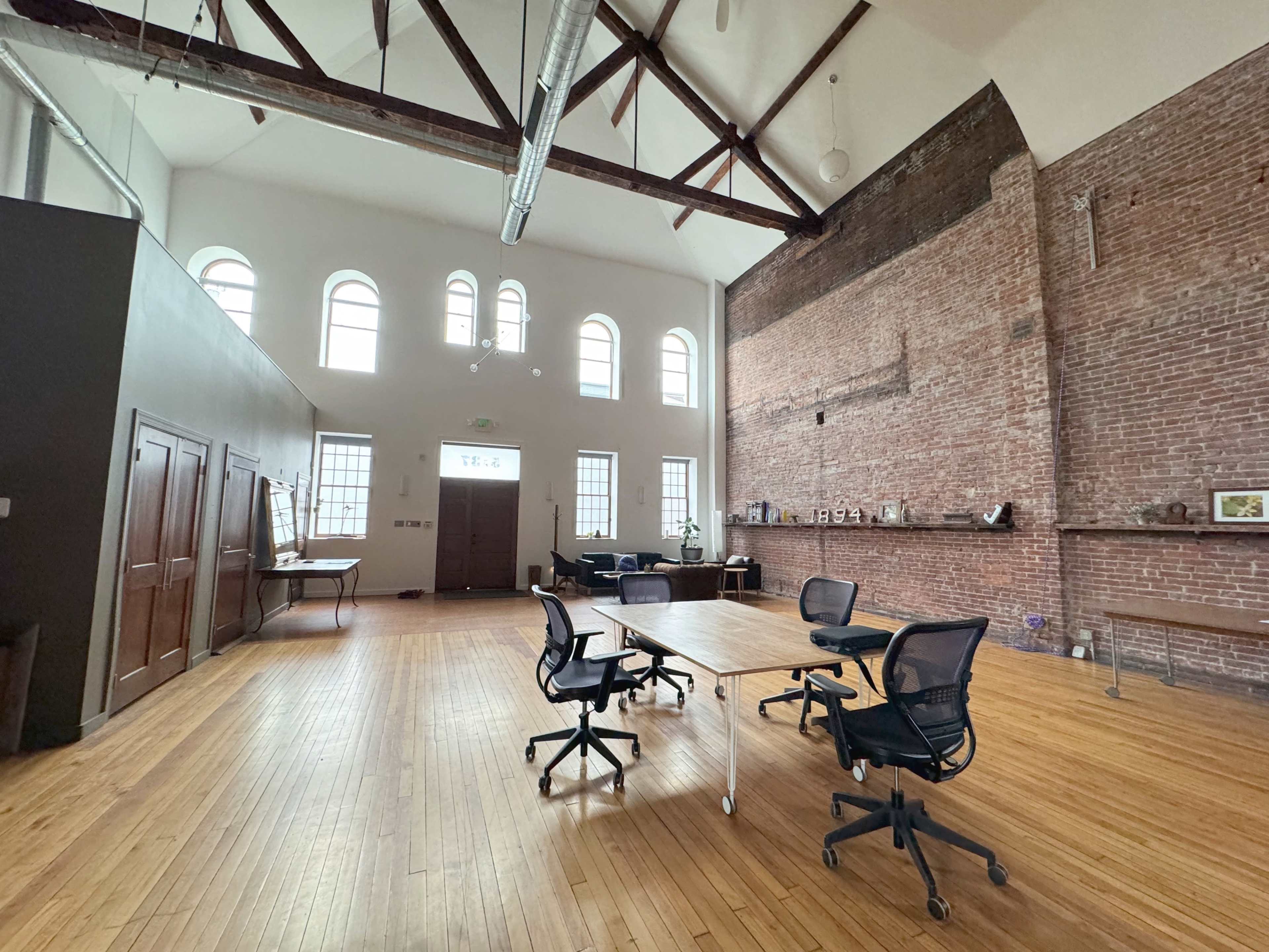 A spacious, modern office with wooden floors, exposed brick walls, and large windows, featuring a table and several rolling chairs.