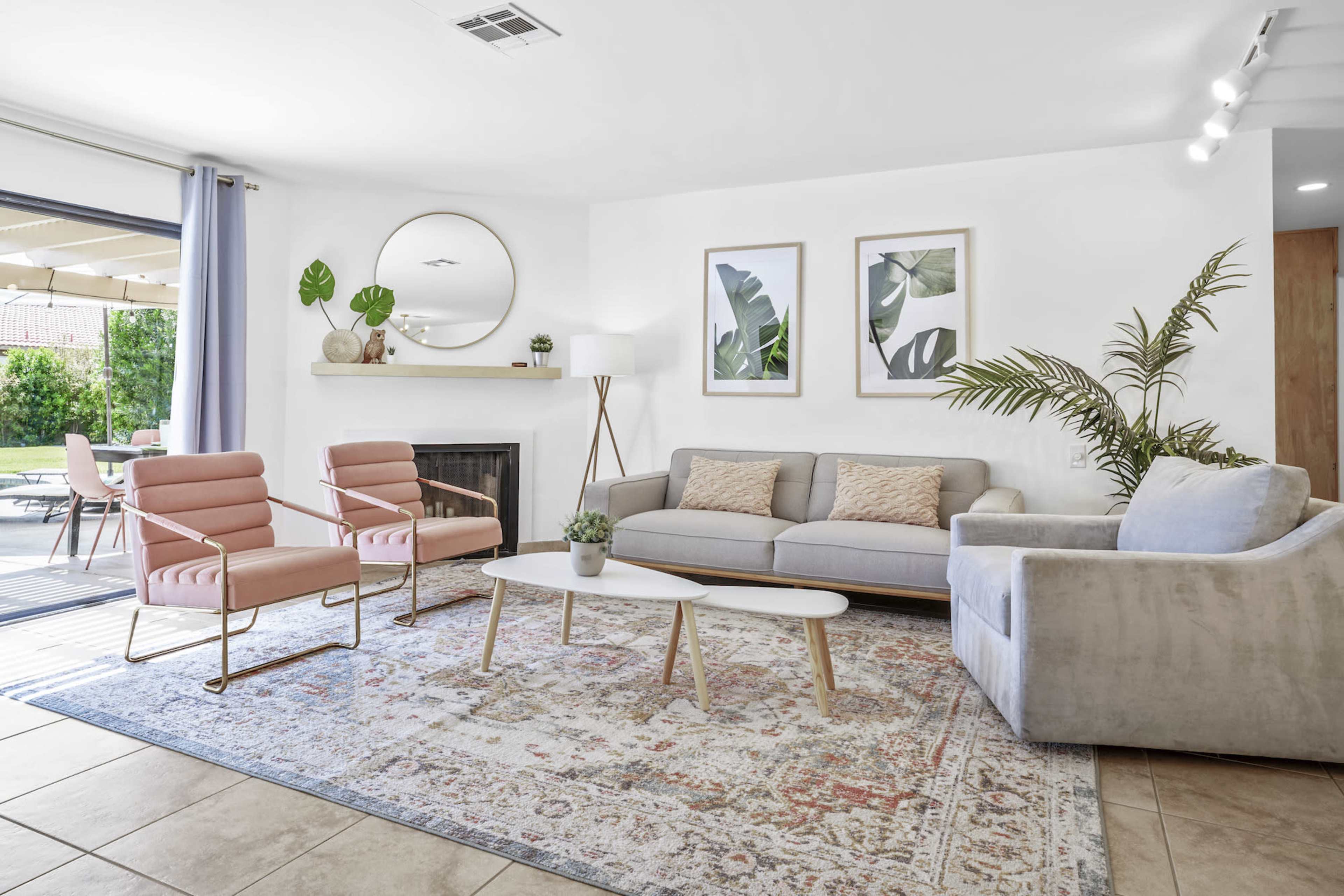 A modern living room featuring a gray sofa, two pink accent chairs, a round mirror, and tropical-themed artwork on the walls, with a sliding door leading to a patio.