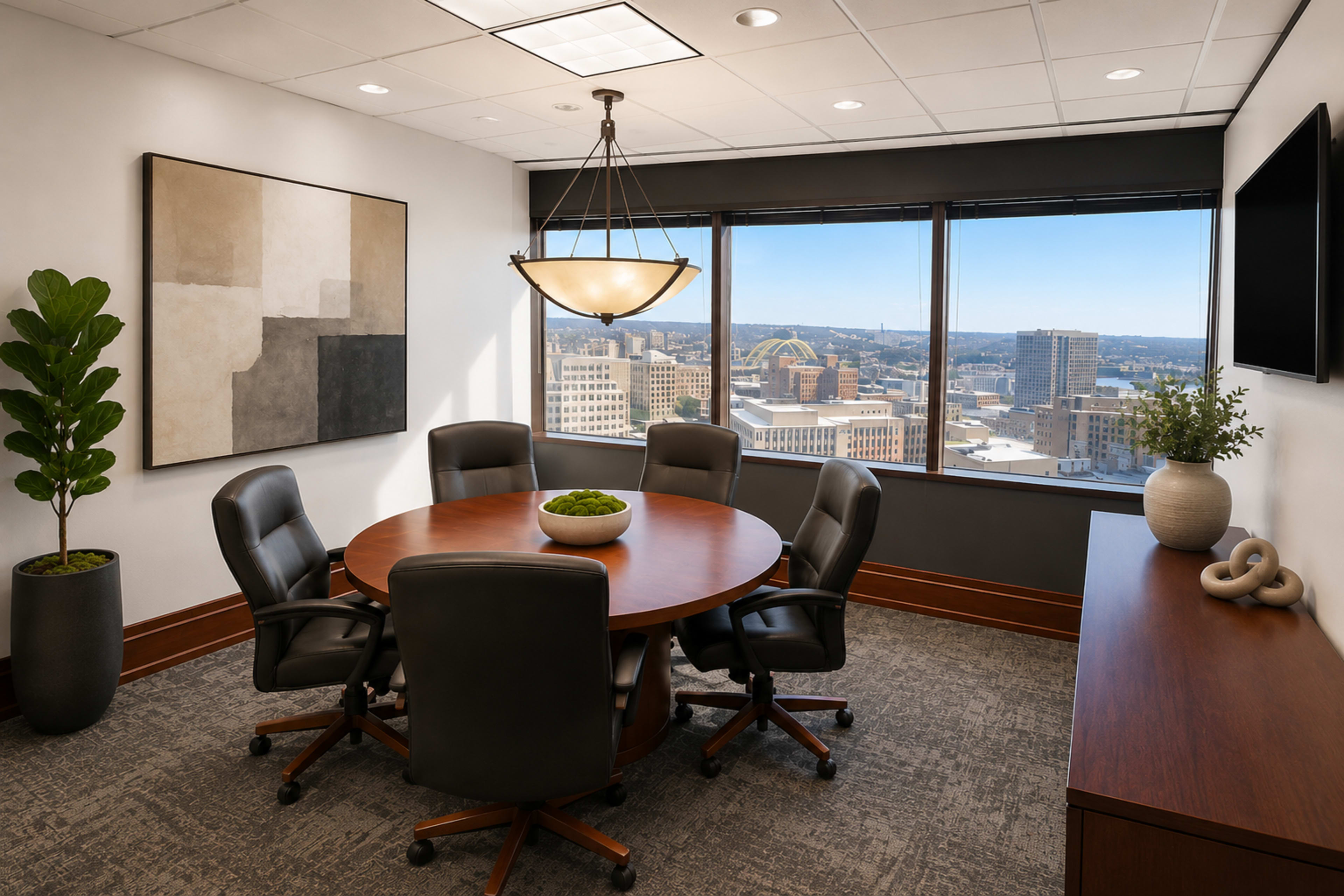 The image shows a modern conference room with a round wooden table surrounded by six black chairs, large windows revealing a cityscape, and minimalistic decor including a plant and artwork.
