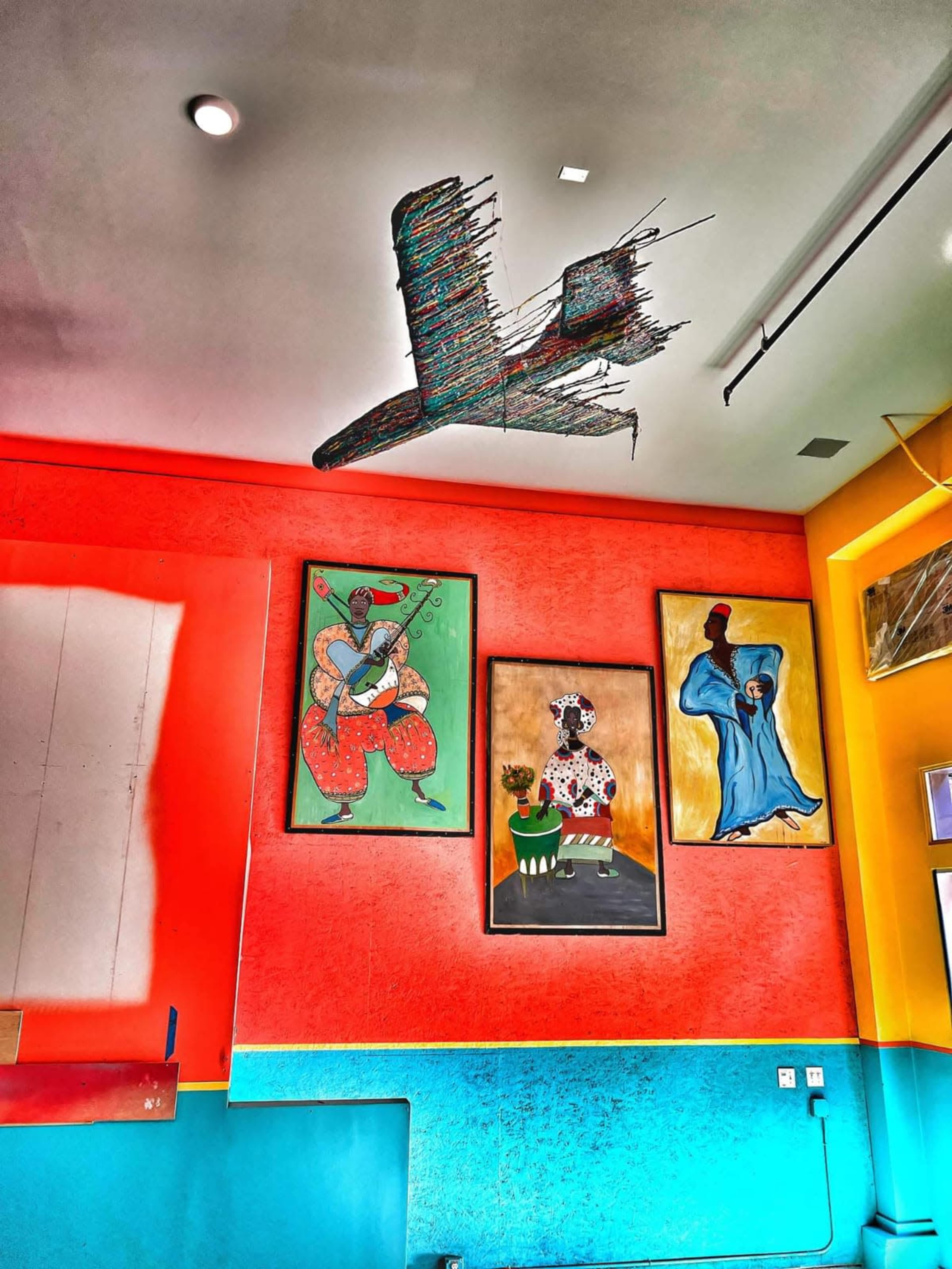 A brightly colored wall featuring three framed paintings and a hanging sculpture resembling an aircraft overhead.