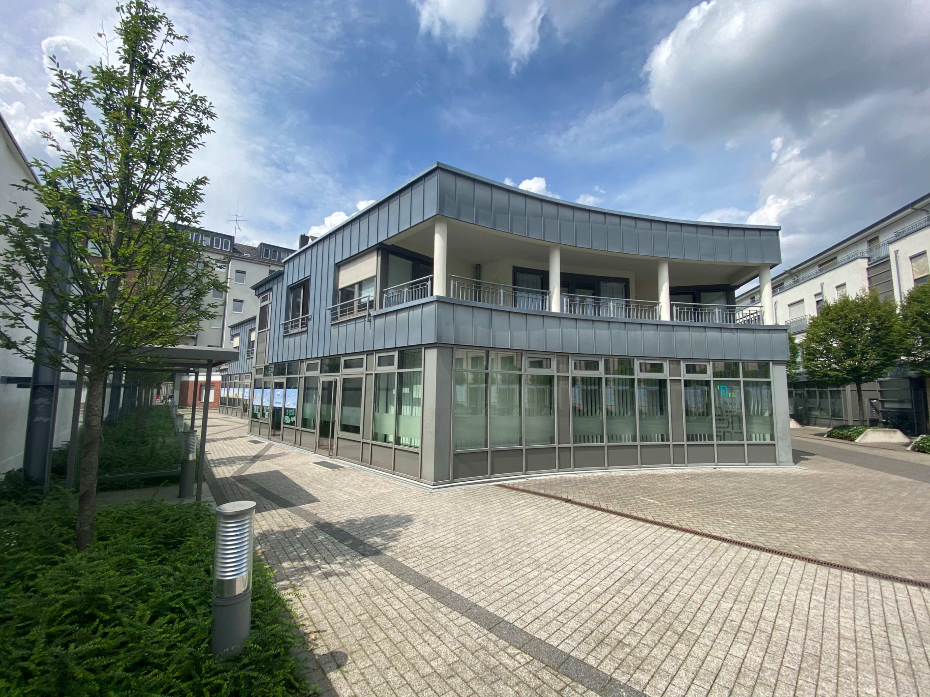 The image shows a modern, curved building with large glass windows, surrounded by a paved courtyard and greenery.