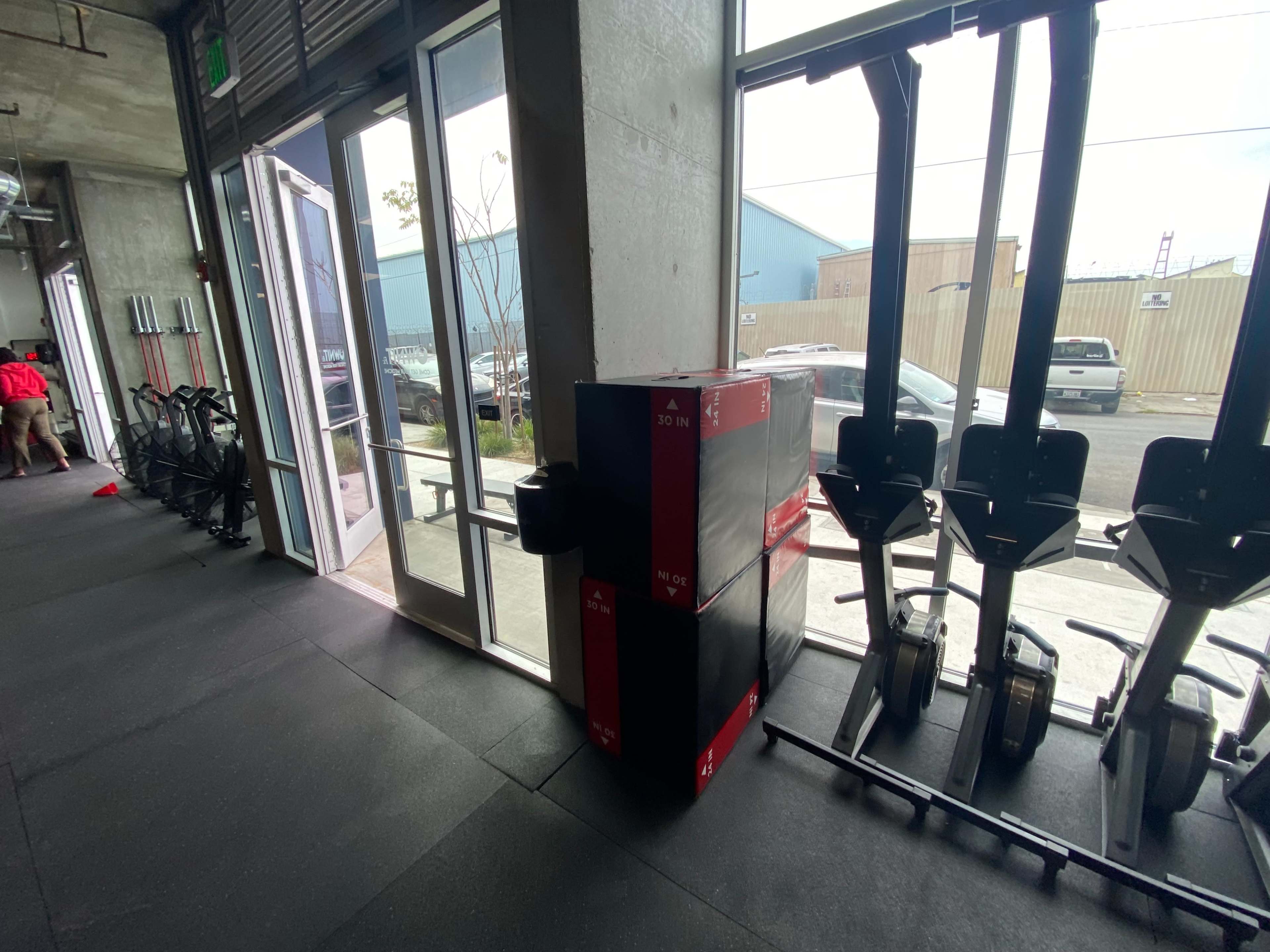 The image shows a corner of a gym with stacked black and red boxes next to exercise equipment and large glass doors leading outside.