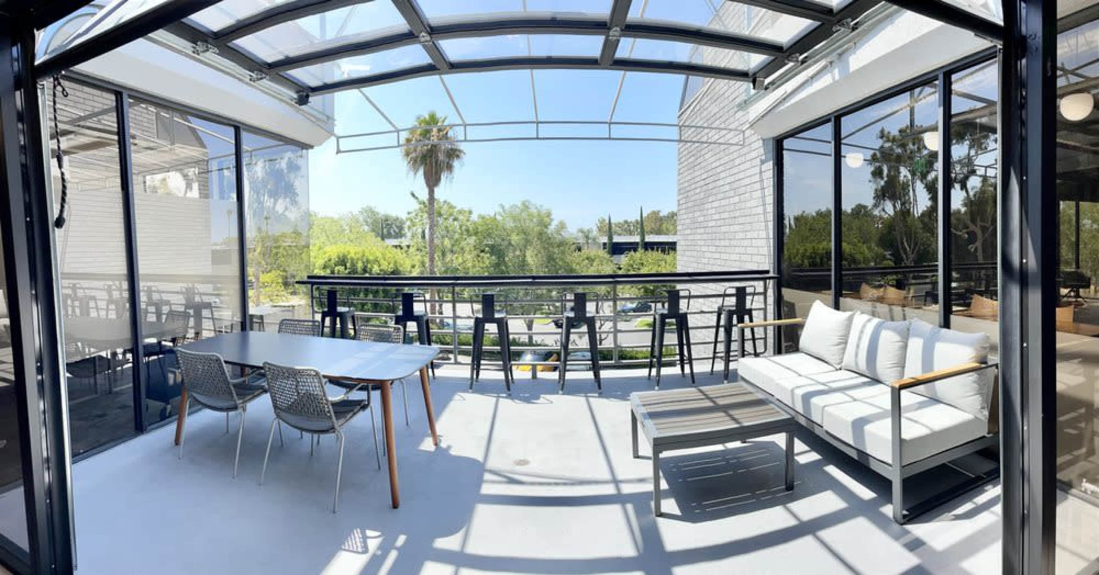 The image shows a spacious outdoor terrace featuring a dining table, chairs, and a lounge area, surrounded by large glass windows and views of greenery.