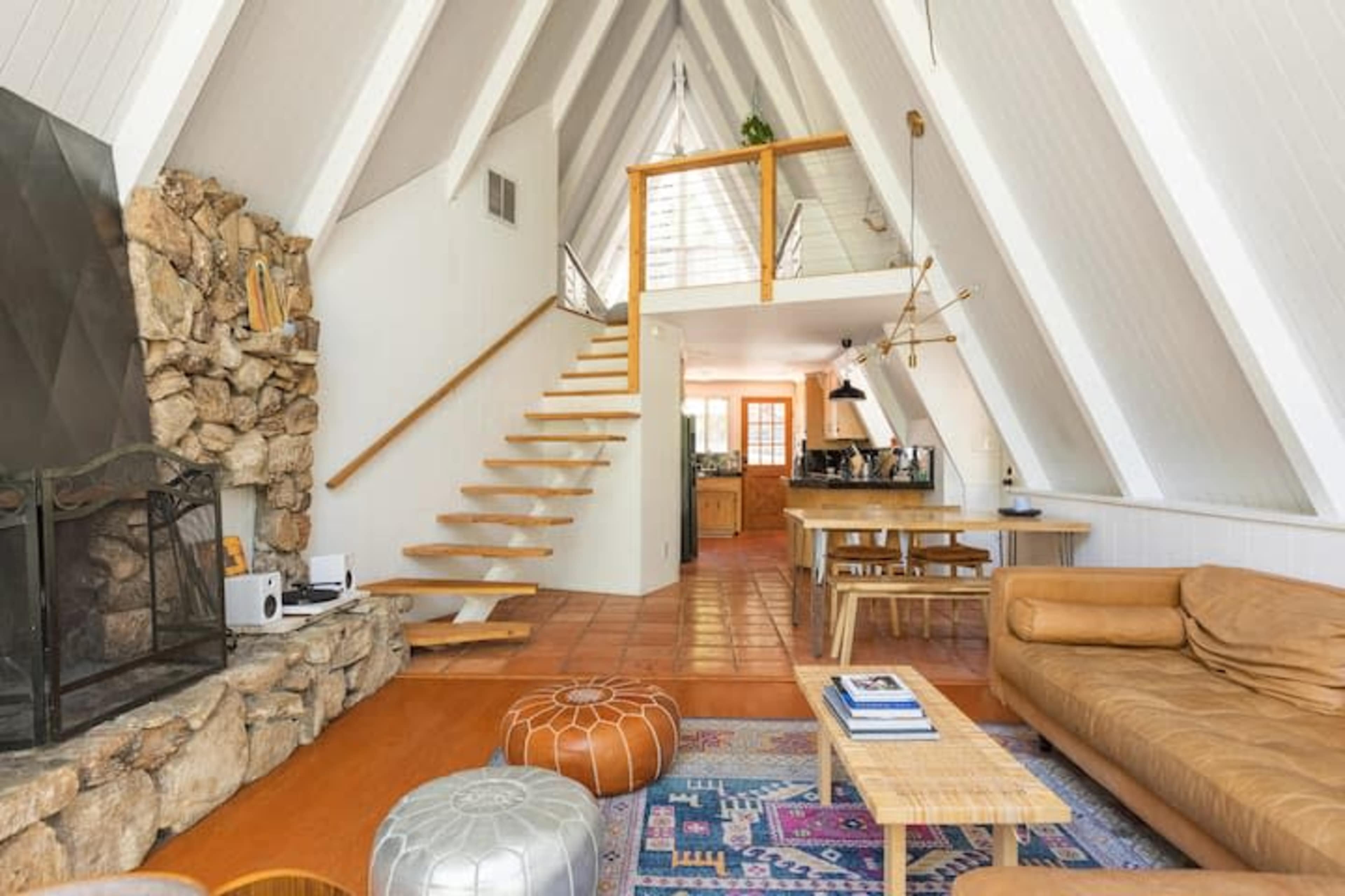 A modern A-frame-style living room featuring a stone fireplace, a staircase leading to a second floor, and an open kitchen in the background.