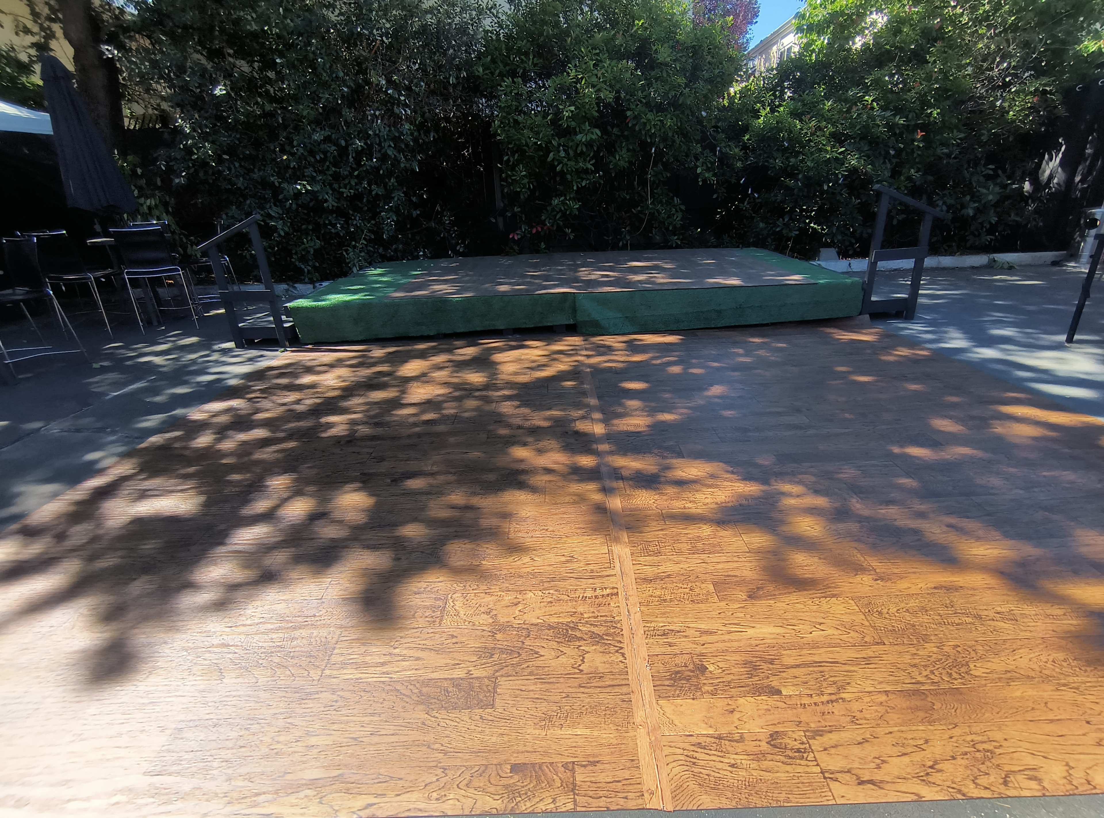 The image shows a wooden stage with a green mat, surrounded by greenery in an outdoor setting.