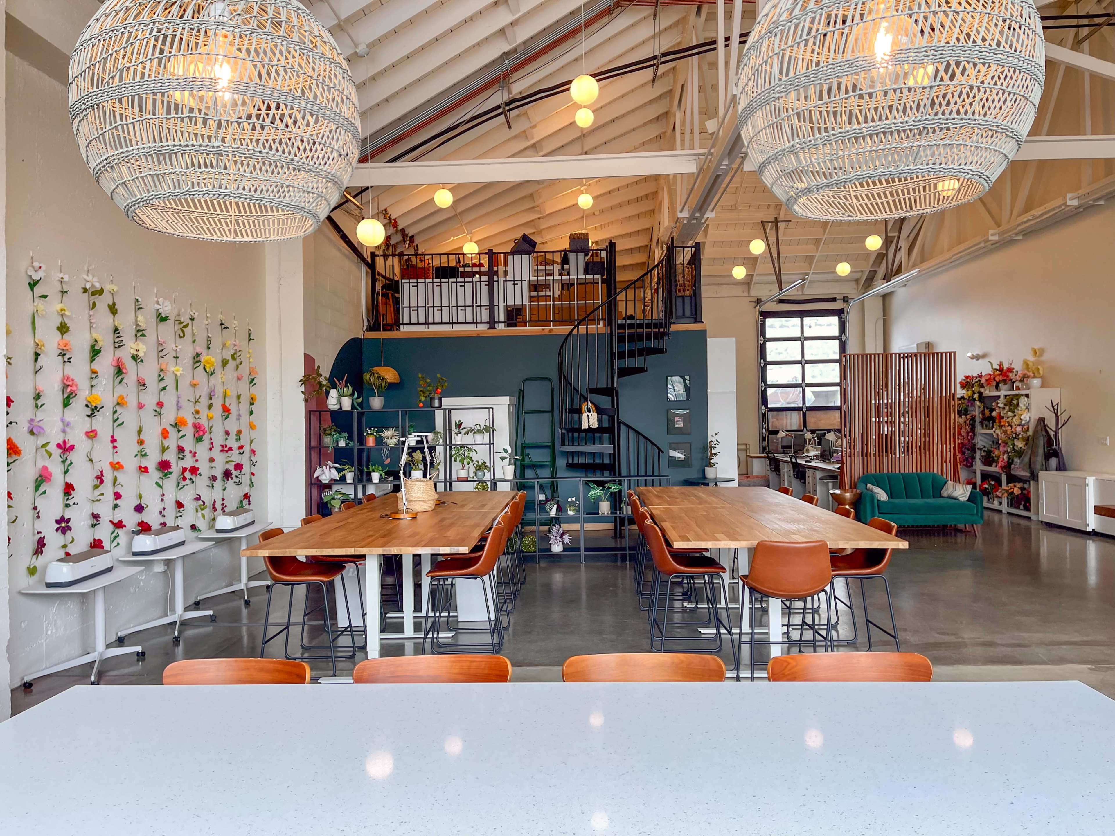The image shows a spacious interior featuring wooden tables, woven pendant lights, and a spiral staircase, with a separation of seating areas and decorative plants on the walls.