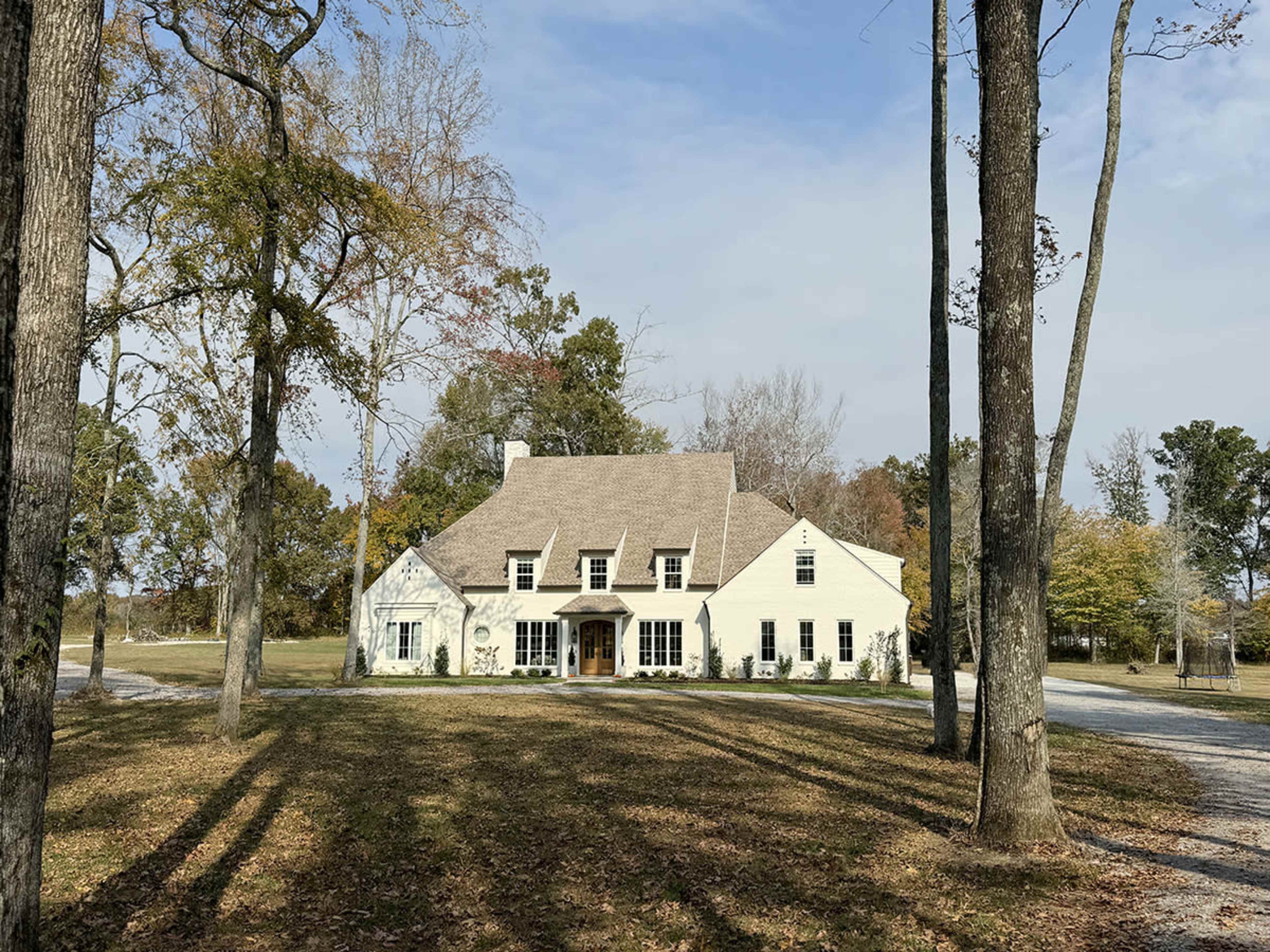 A large, light-colored house with a steeply pitched roof is surrounded by trees and a grassy area.