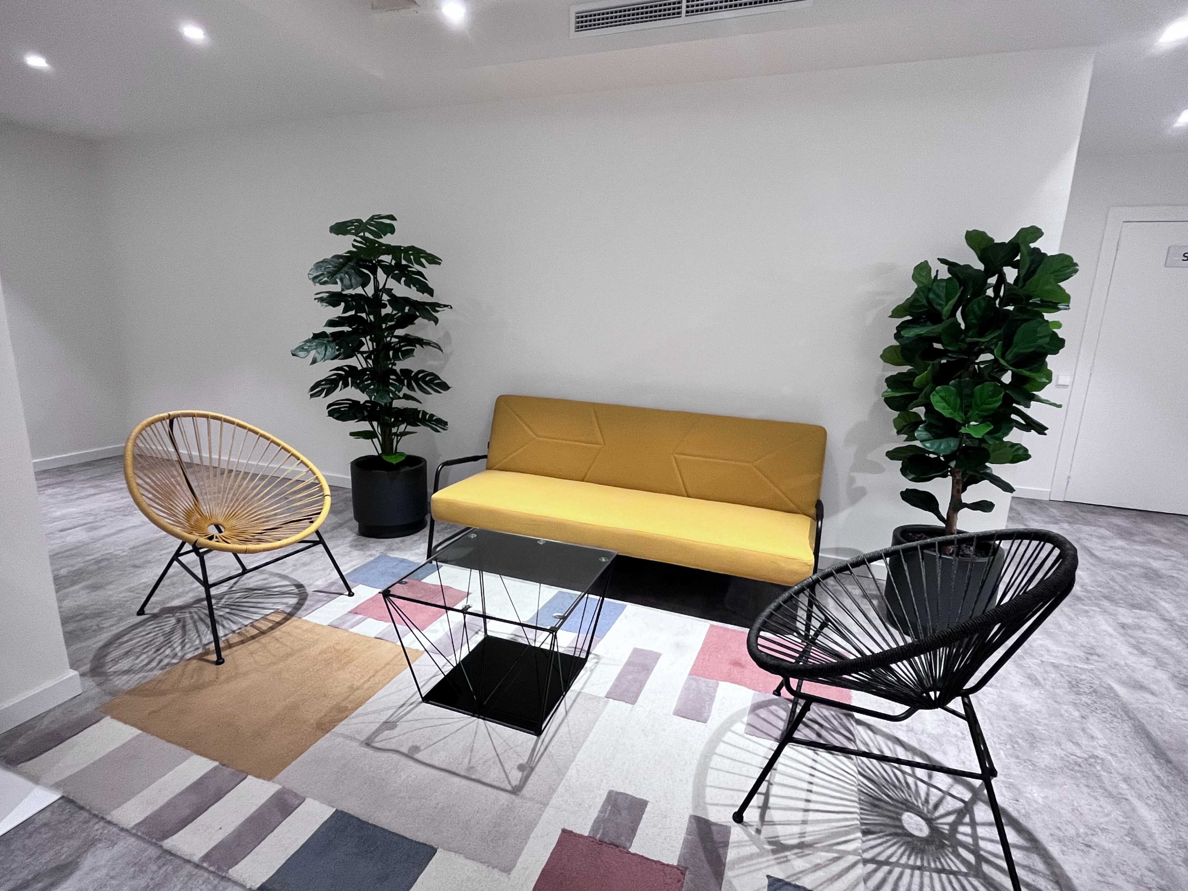 The image shows a modern lounge area with a yellow sofa, a glass coffee table, and two black wire chairs, all positioned on a multicolored rug.
