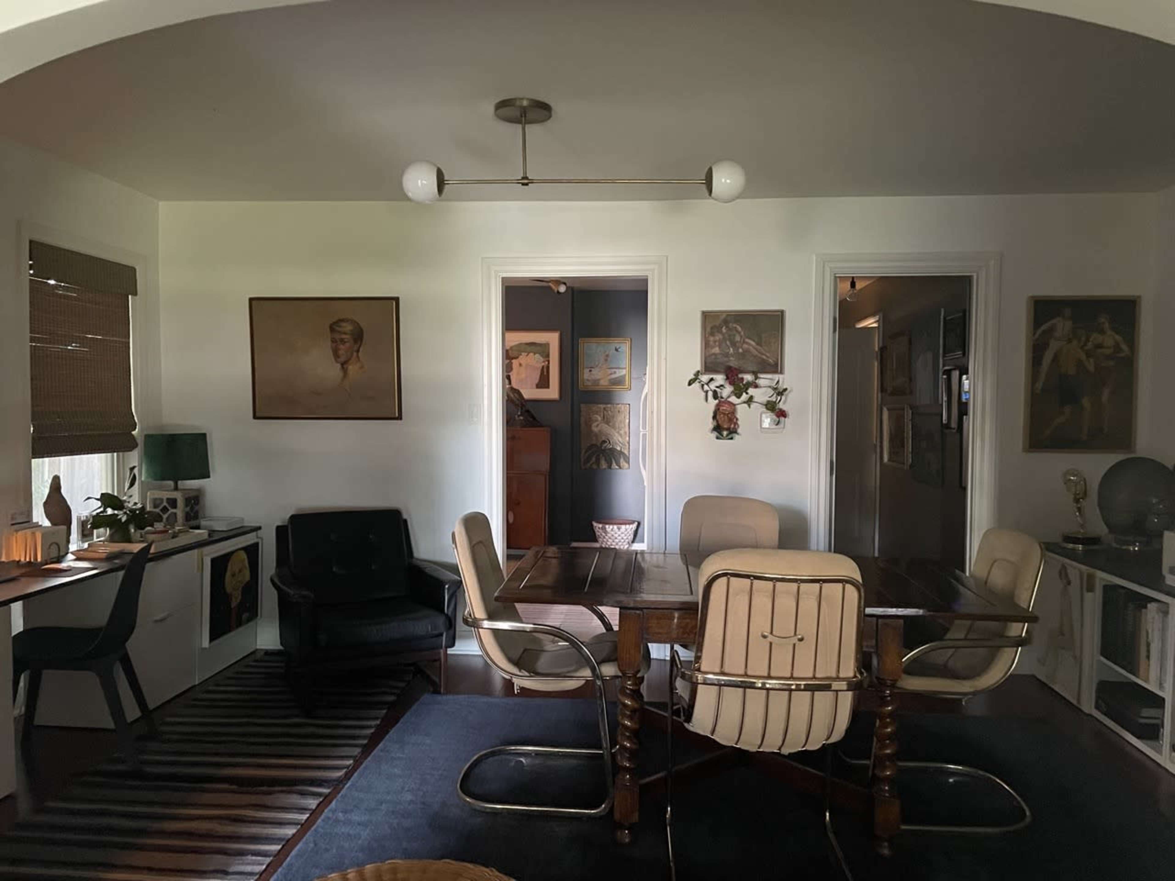 The image shows a dining area with a wooden table surrounded by chairs, framed artwork on the walls, and a small desk in the corner.