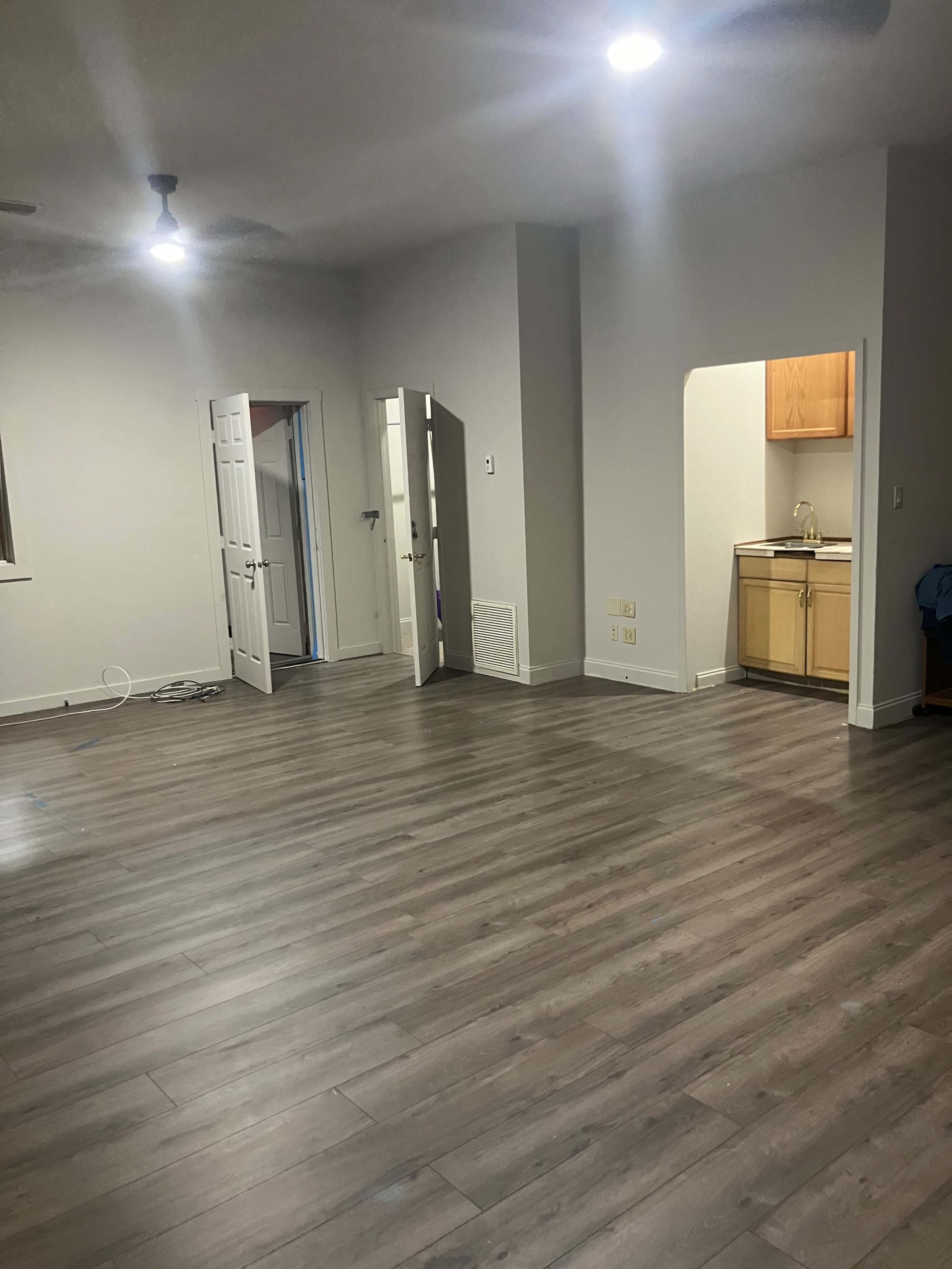 The image shows a spacious room with wooden flooring, two open doors leading to separate areas, and a kitchenette visible in an alcove.