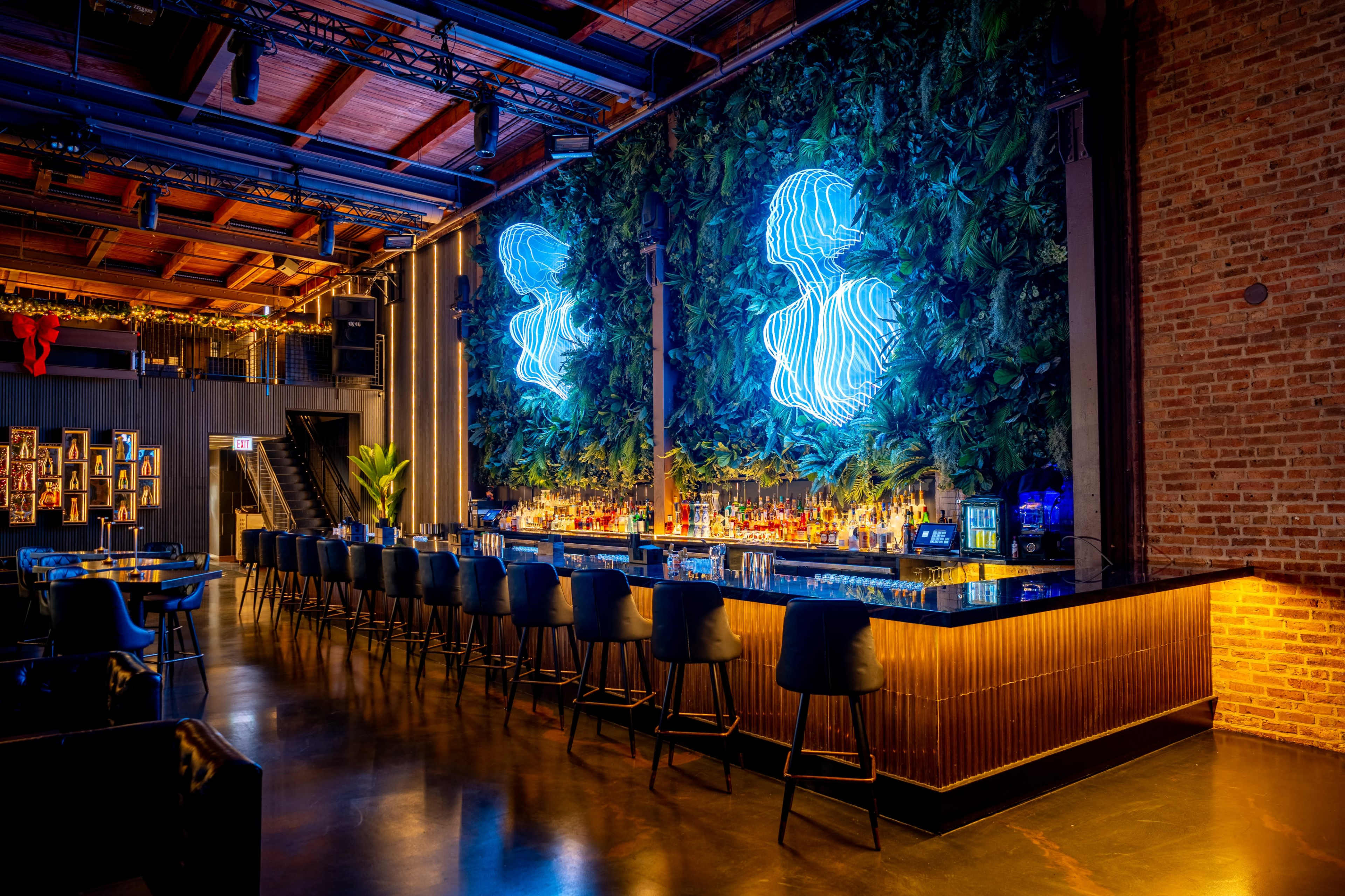 The image shows a modern bar with a neon wall display and a lush greenery backdrop, featuring a long counter lined with high-backed chairs.