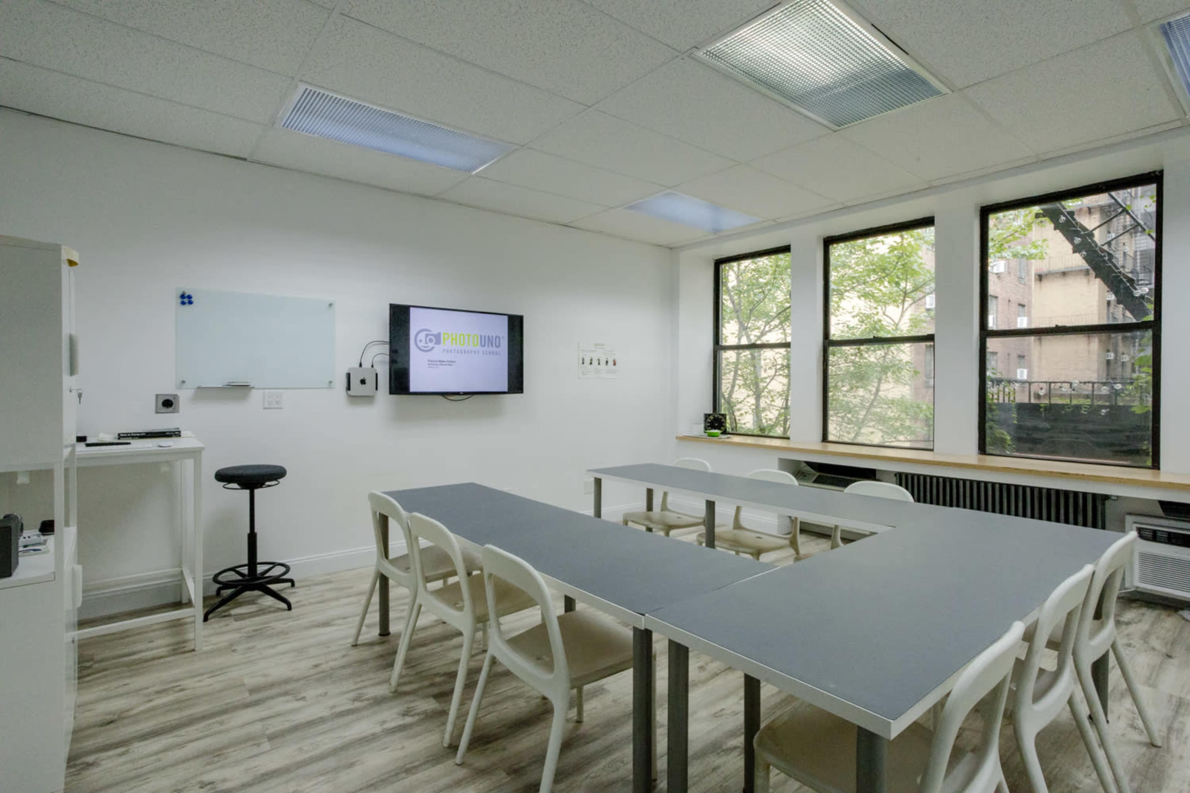The image shows a modern classroom with multiple tables arranged in a U-shape, a presentation screen on the wall, and large windows allowing natural light.