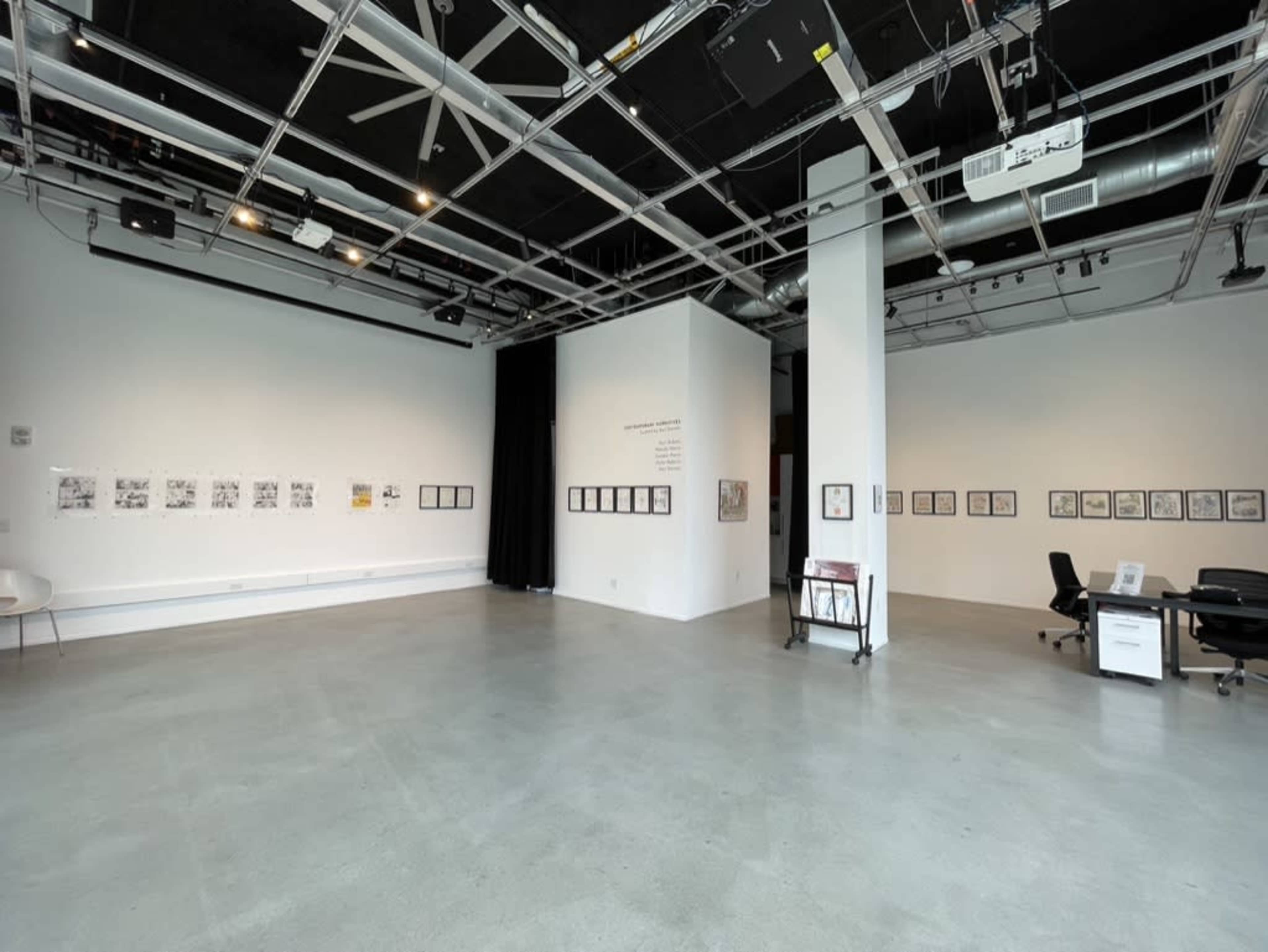 The image shows a spacious art gallery featuring white walls, a concrete floor, and several framed artworks displayed on the walls.