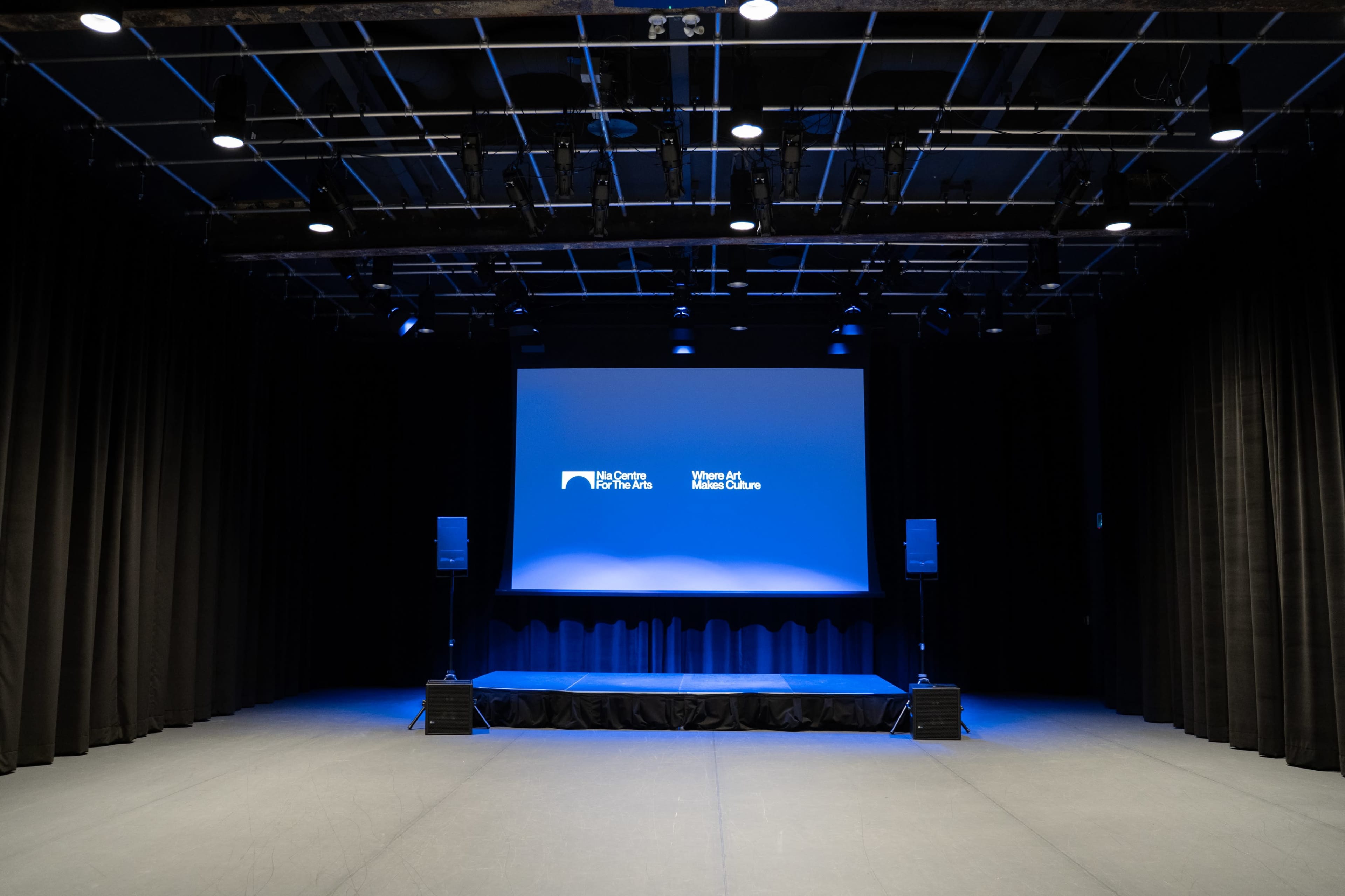 The image shows an empty stage with a large screen and two speakers on either side, set against a backdrop of dark curtains.