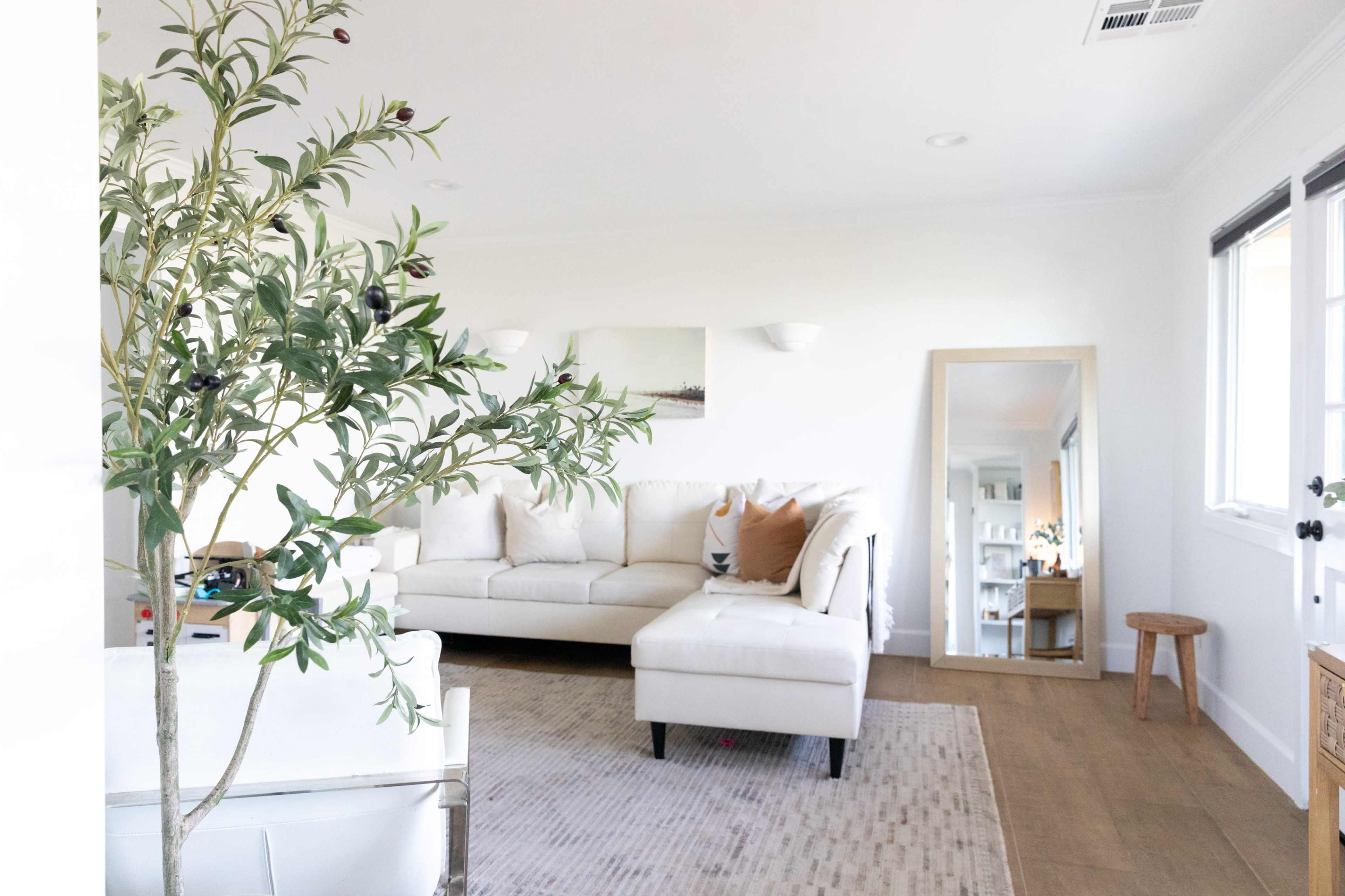 The image shows a bright living room featuring a white sectional sofa, a large mirror, a small wooden stool, and a decorative potted plant.