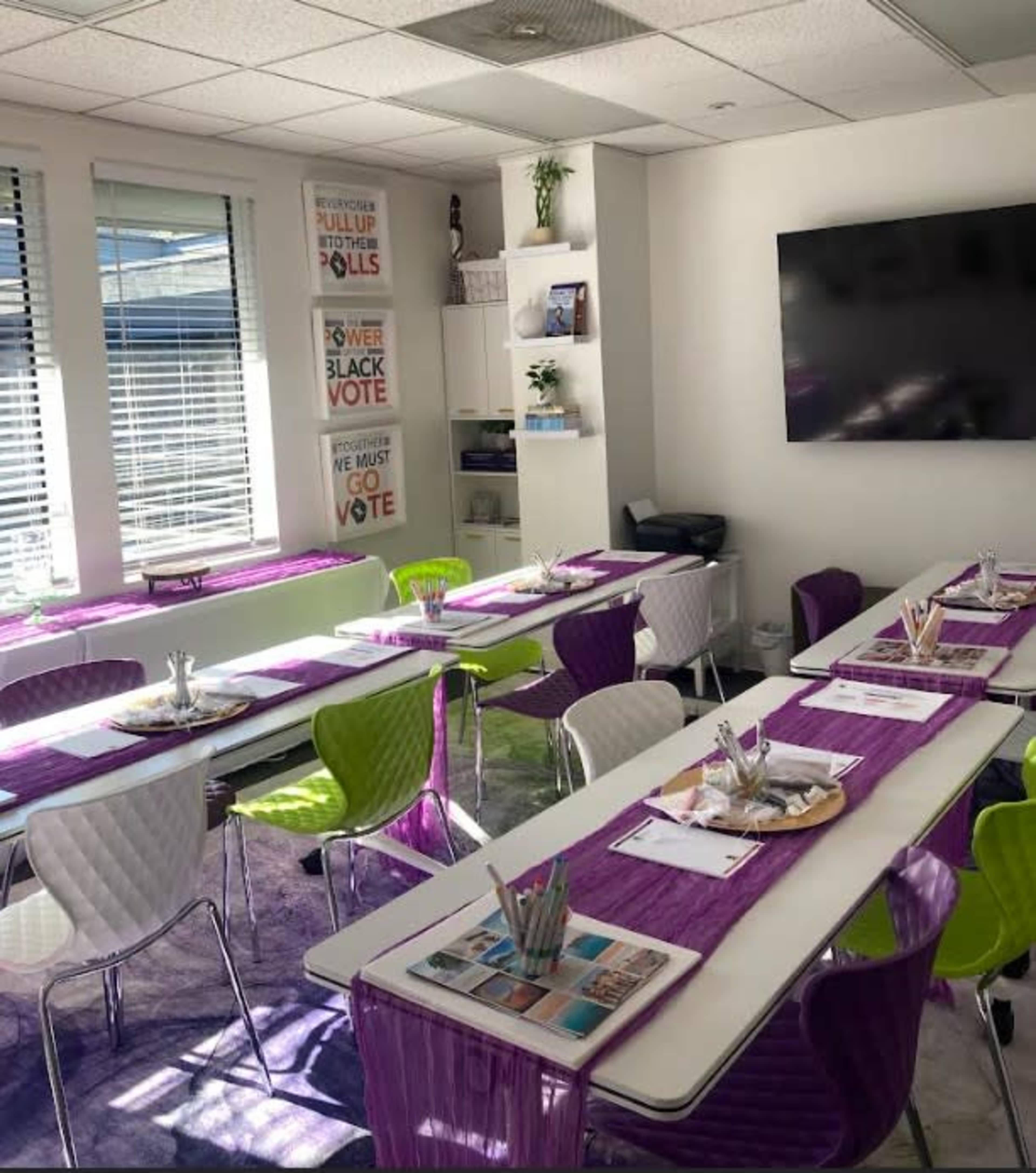 The image shows a brightly lit conference room with tables set for a meeting, featuring chairs in white and green, and decorated with purple table runners and stationery.