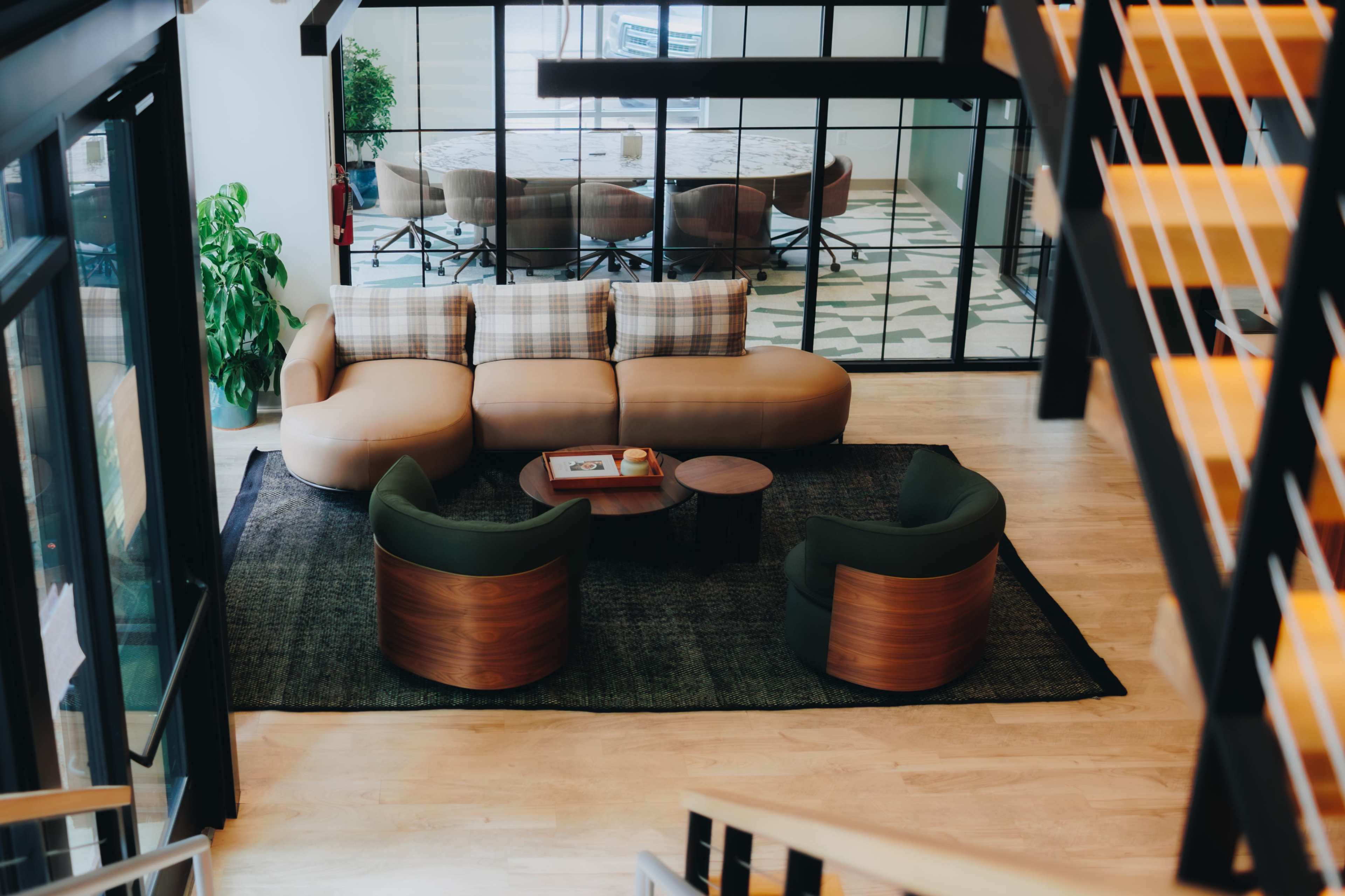 The image shows a modern lounge area with a curved sofa and two circular chairs arranged around a small table on a dark rug, with a large glass-walled meeting room visible in the background.
