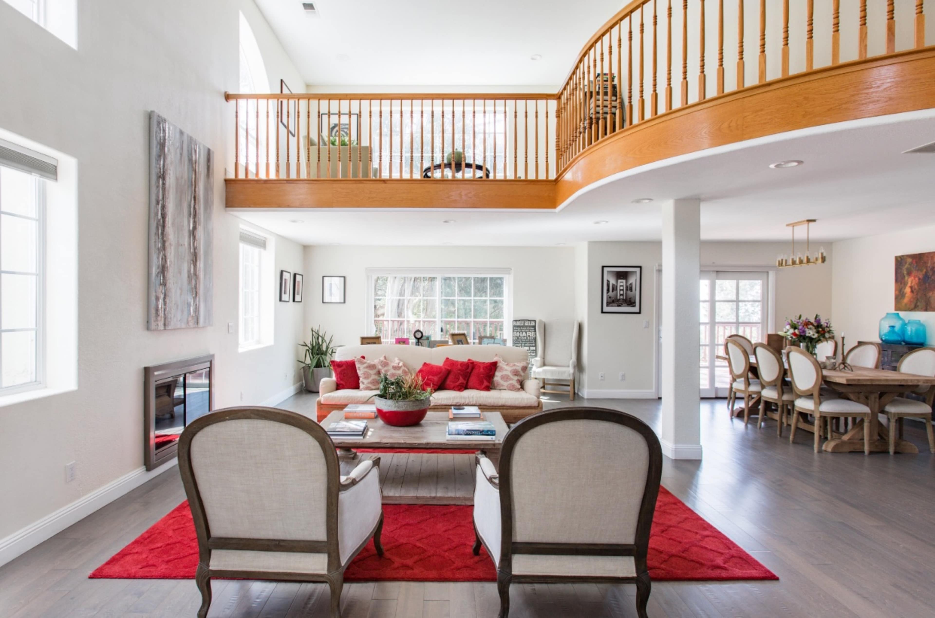 A spacious, well-lit living area featuring a seating arrangement with two chairs, a sofa adorned with red cushions, and an upper balcony overlooking the space.