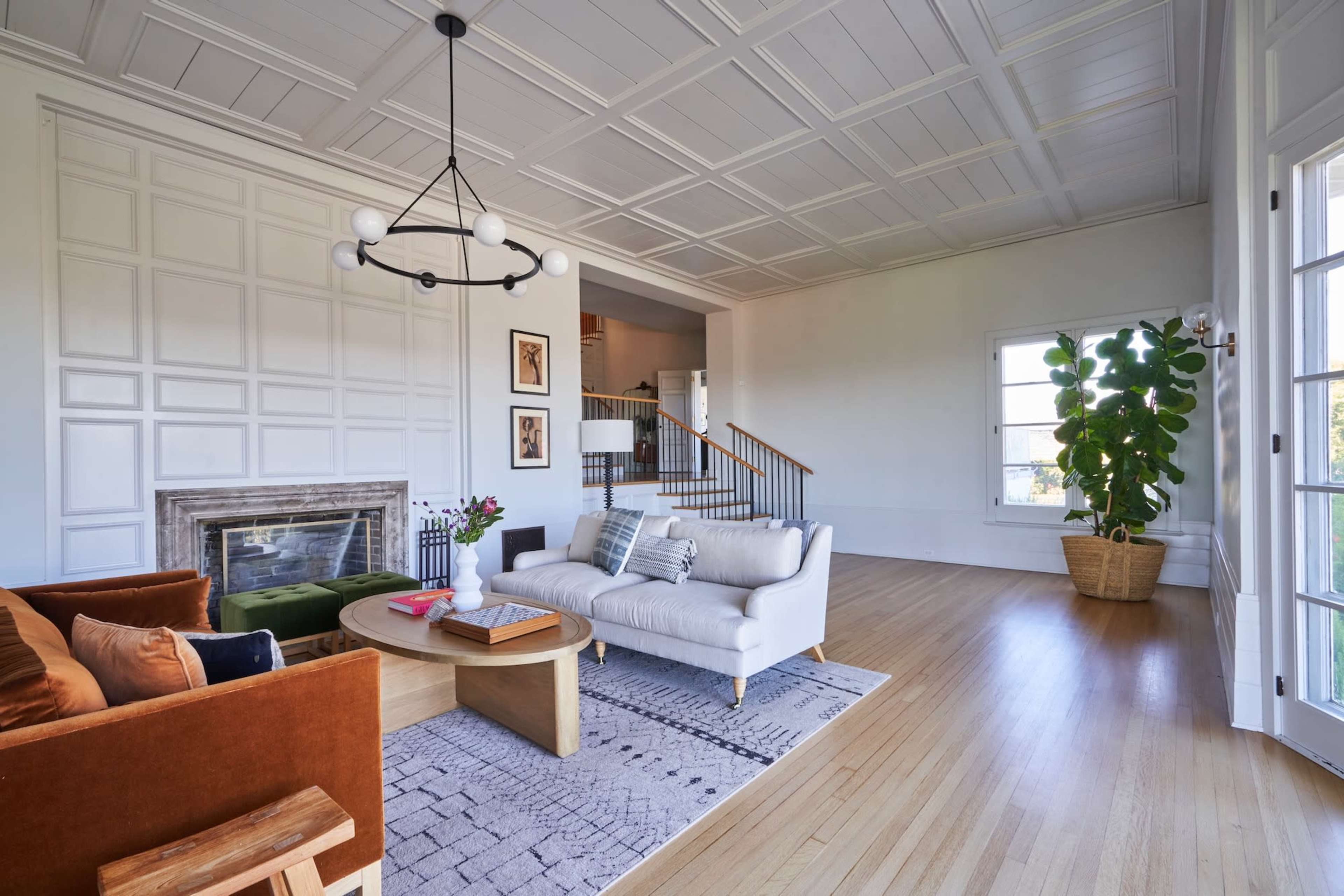 The image shows a modern living room with a white sofa, an orange armchair, a round coffee table, a fireplace, and large windows that allow natural light in.
