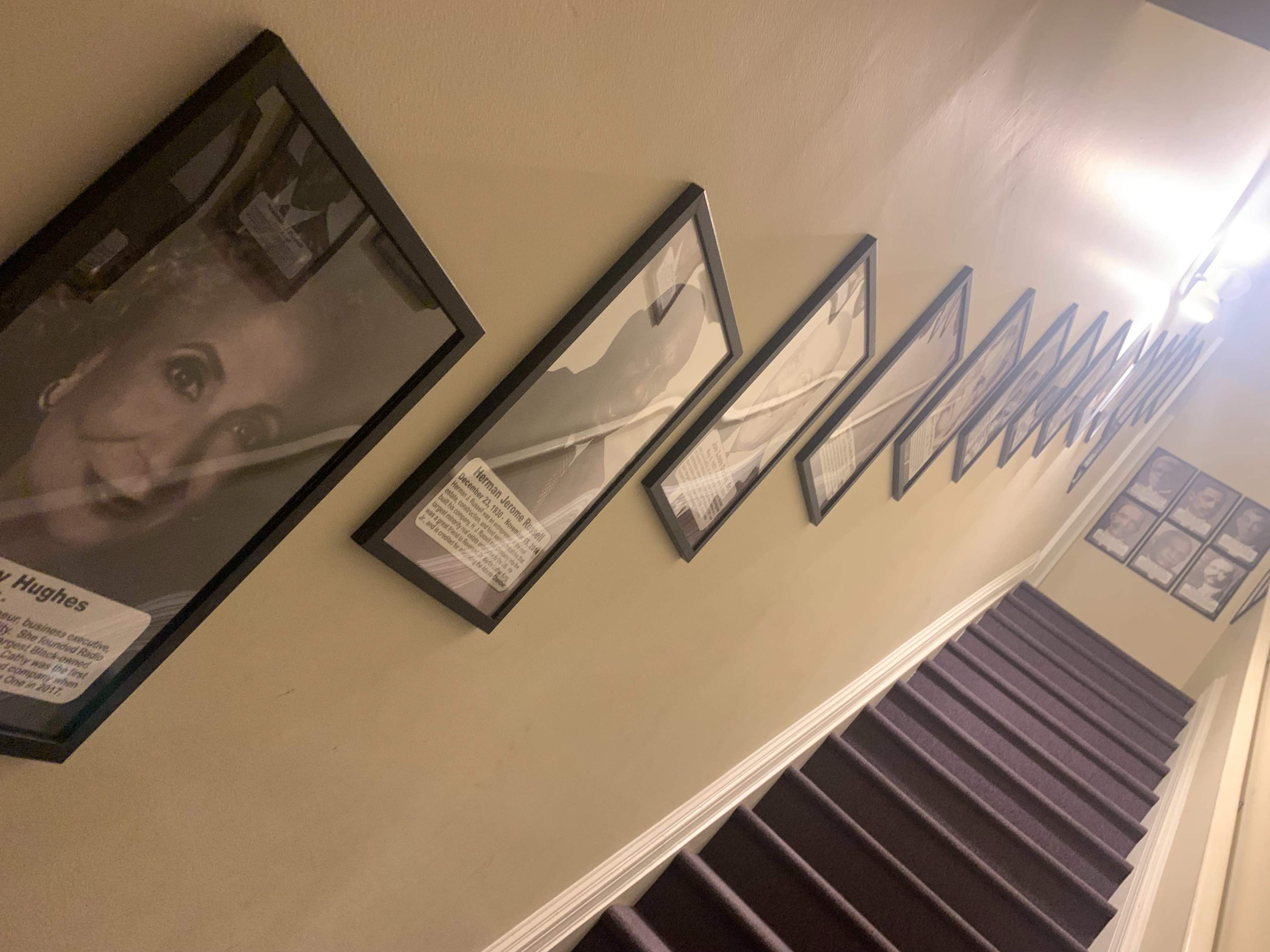 The image shows a stairway with a series of framed photographs and information displayed on the walls, leading upward.