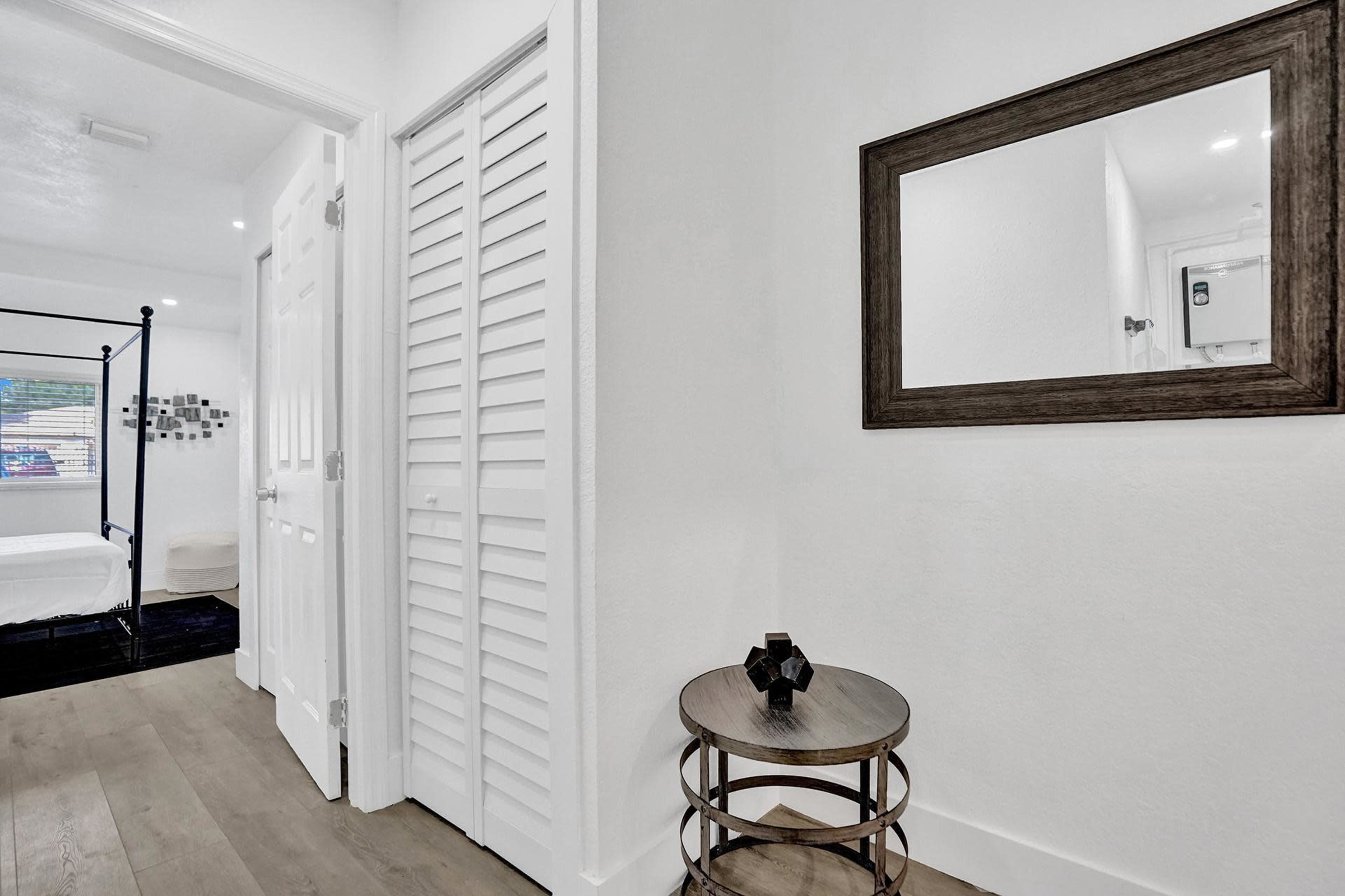 A hallway features a round side table, a mirror on the wall, and a closed closet with louvered doors, leading to a bedroom.