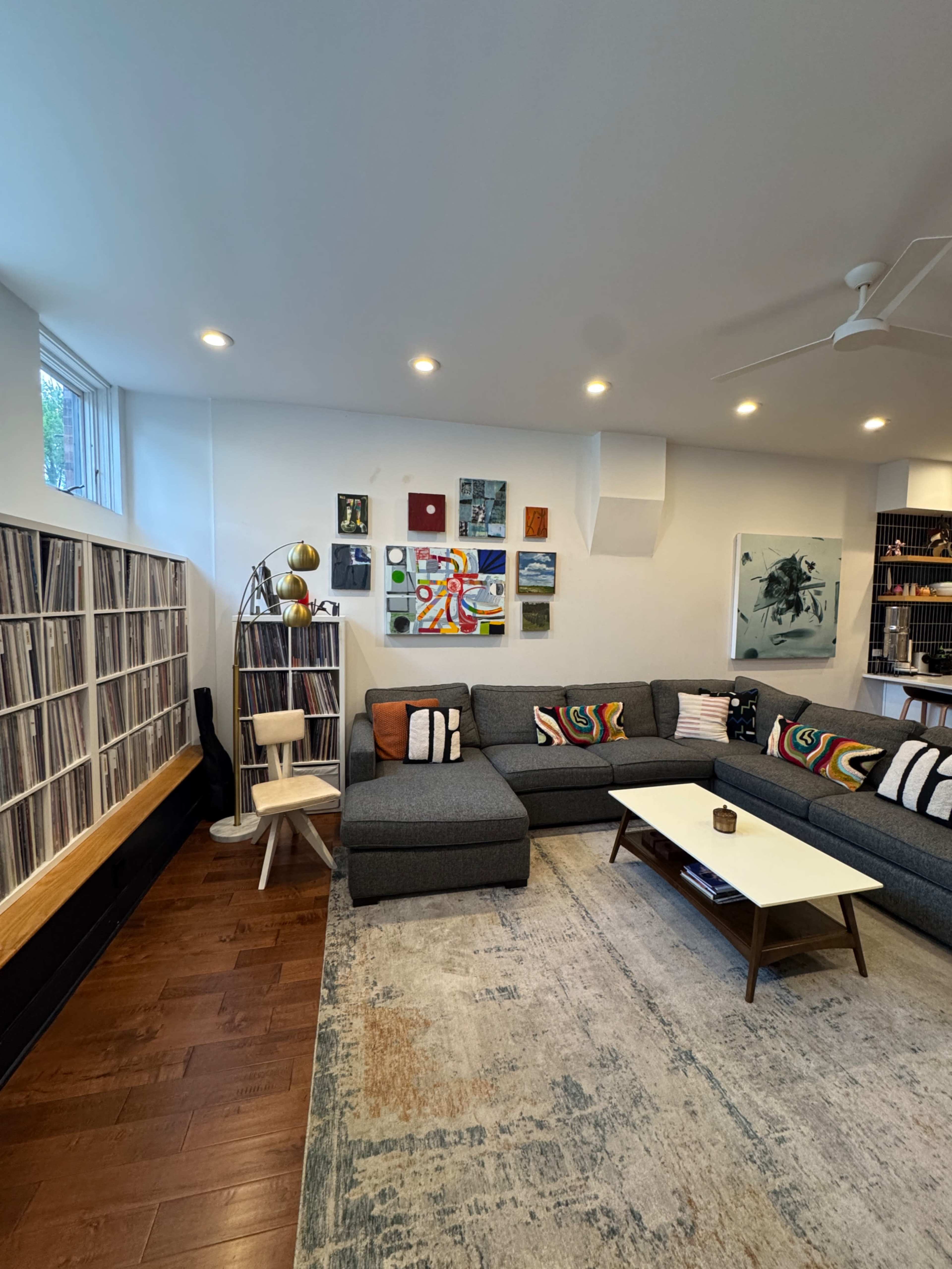The image shows a modern living room with a gray sectional sofa, a coffee table, a large area rug, and a wall of vinyl records.