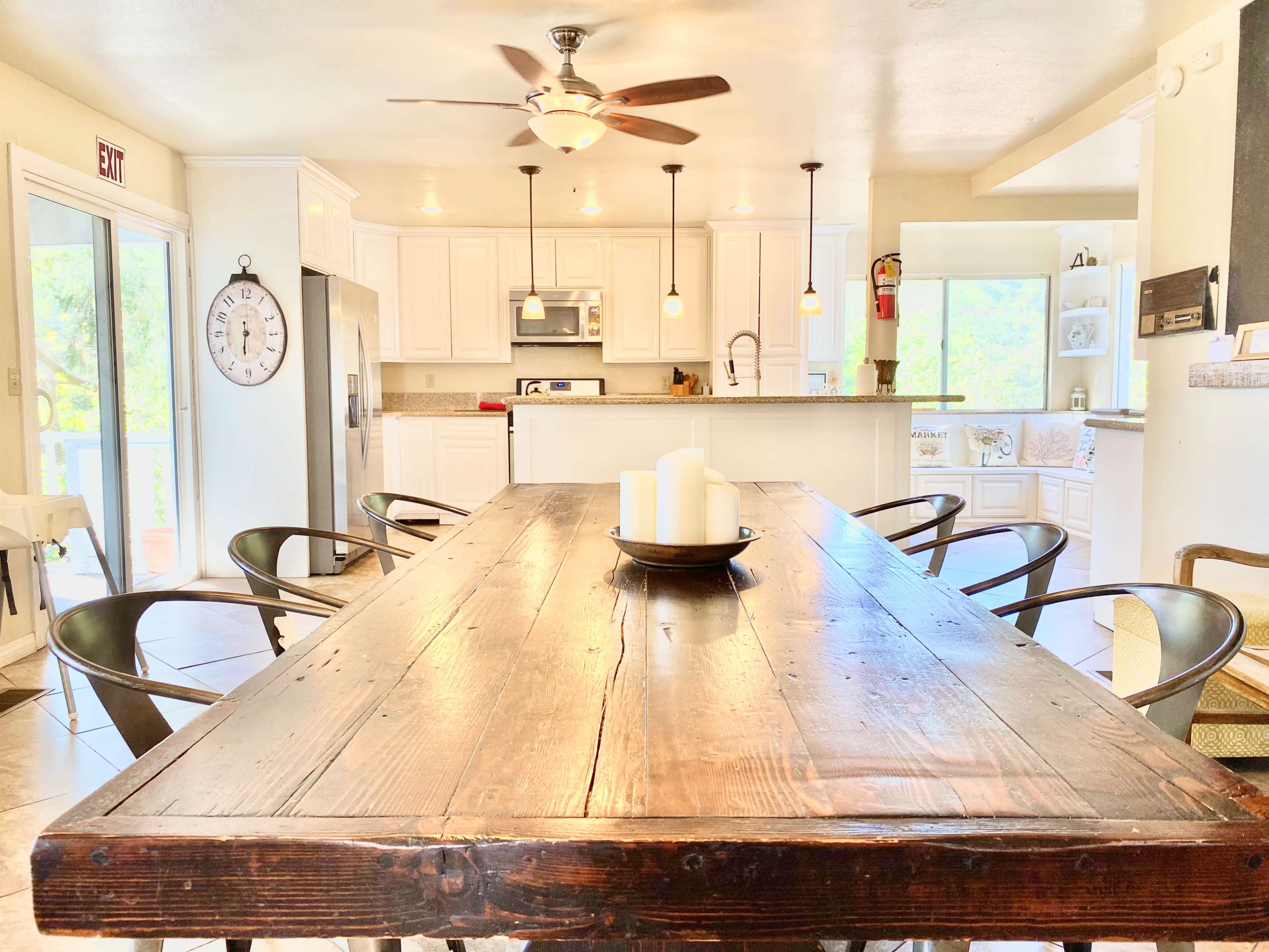 The image shows a bright kitchen-dining area with a large wooden table in the foreground and white cabinetry in the background.