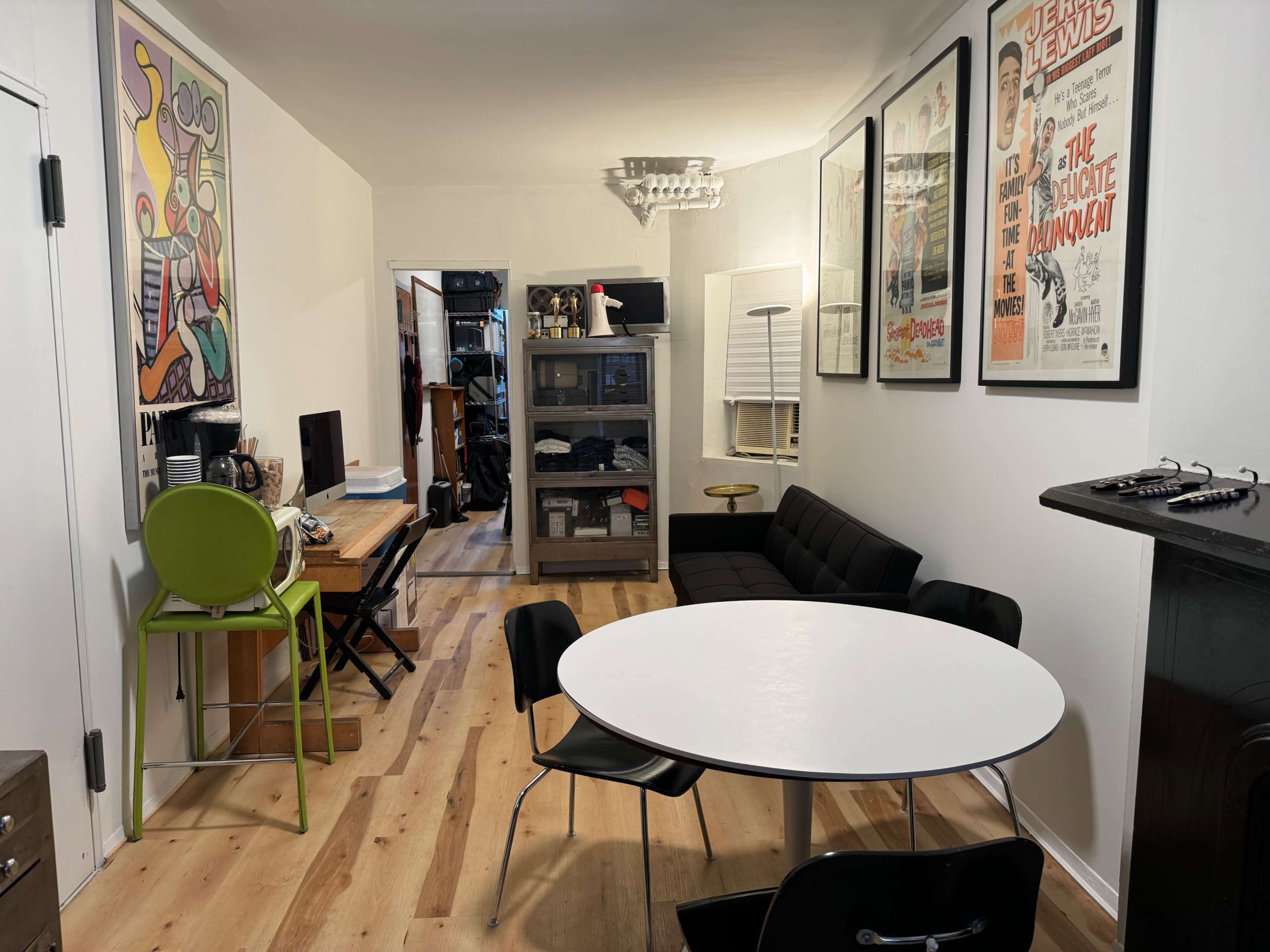 The image shows a small, modern living space with a round white table, black chairs, a black sofa, a wooden desk, and several framed posters on the walls.