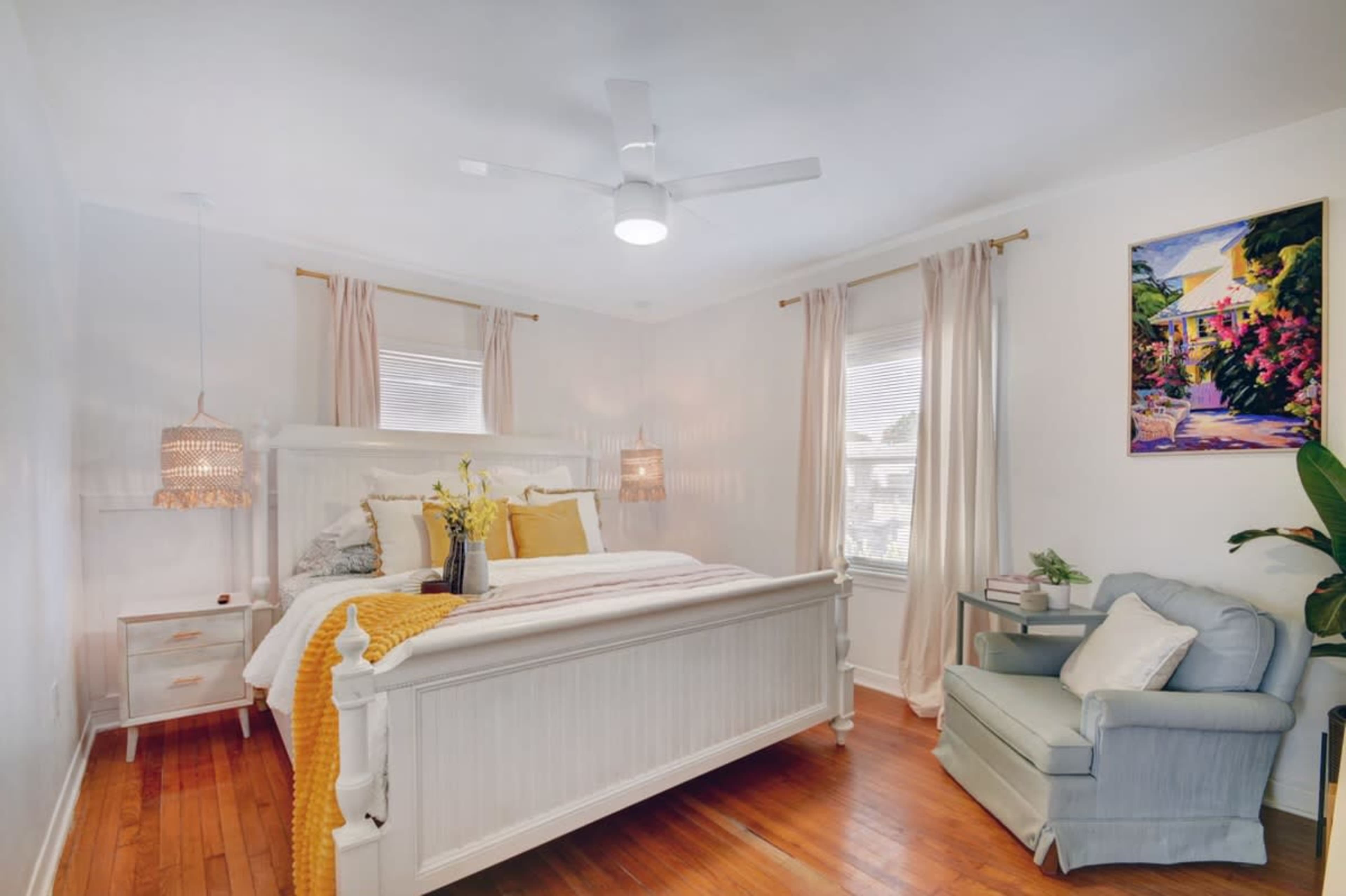A bright bedroom features a large bed with yellow and white bedding, a blue armchair, and decorative wall art.