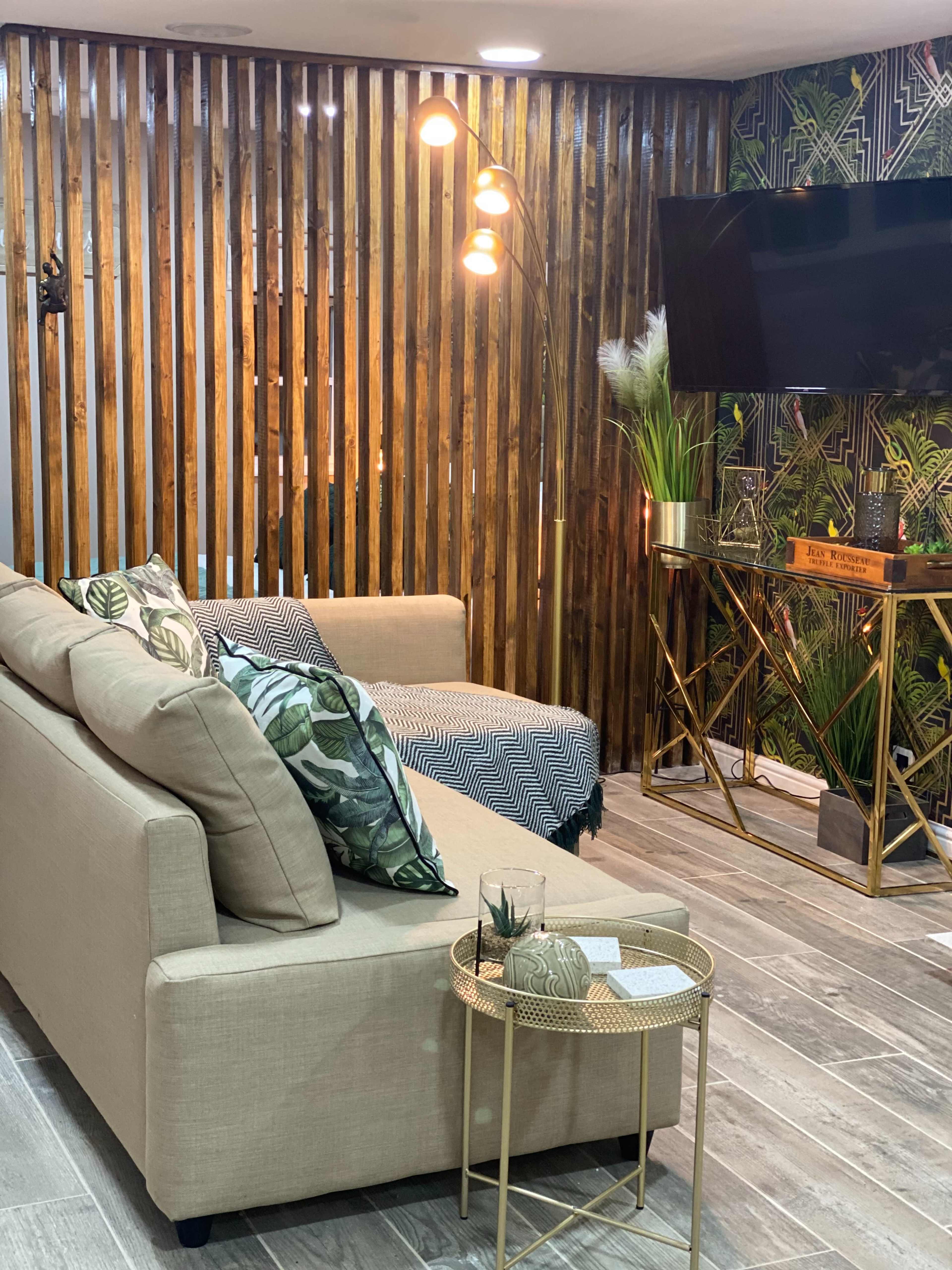 A modern living room with a beige sofa, decorative cushions, a floor lamp, and a wall adorned with wooden slats and patterned wallpaper.